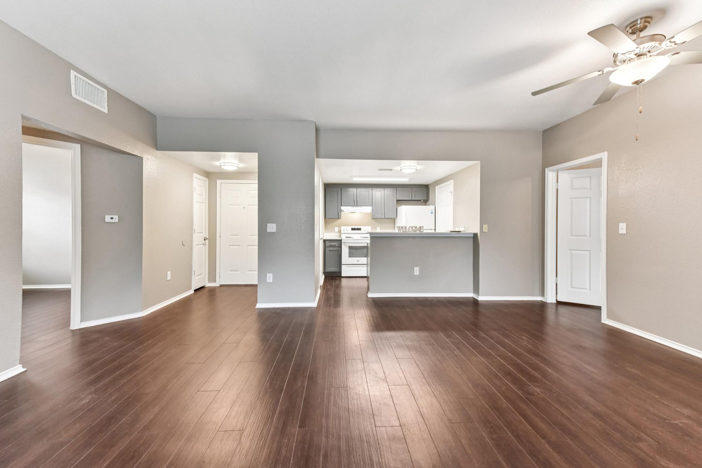 Spacious living area with hardwood floors, featuring a ceiling fan and open layout. The kitchen is visible in the background, showcasing modern appliances and cabinetry. Two doorways lead to separate rooms, enhancing the flow of the space. The walls are painted in a light gray color, creating a bright atmosphere.