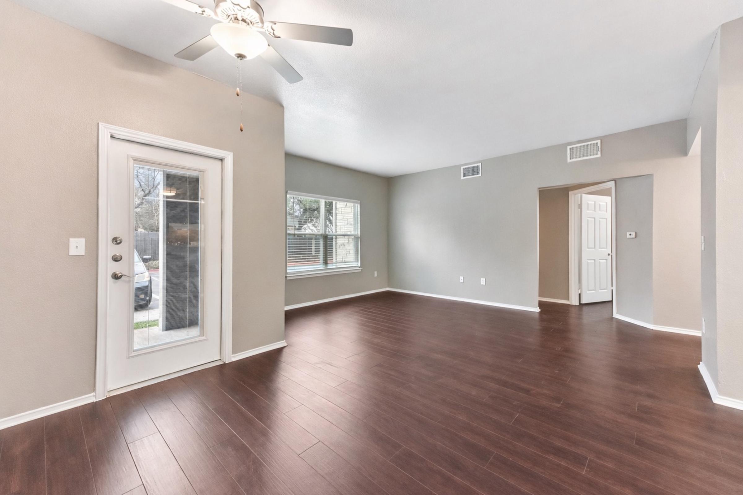 Spacious living room with hardwood flooring, ceiling fan, and large windows letting in natural light. Features a sliding glass door leading to an outdoor area and an entrance to another room on the right. Neutral-colored walls enhance the airy feel of the space.