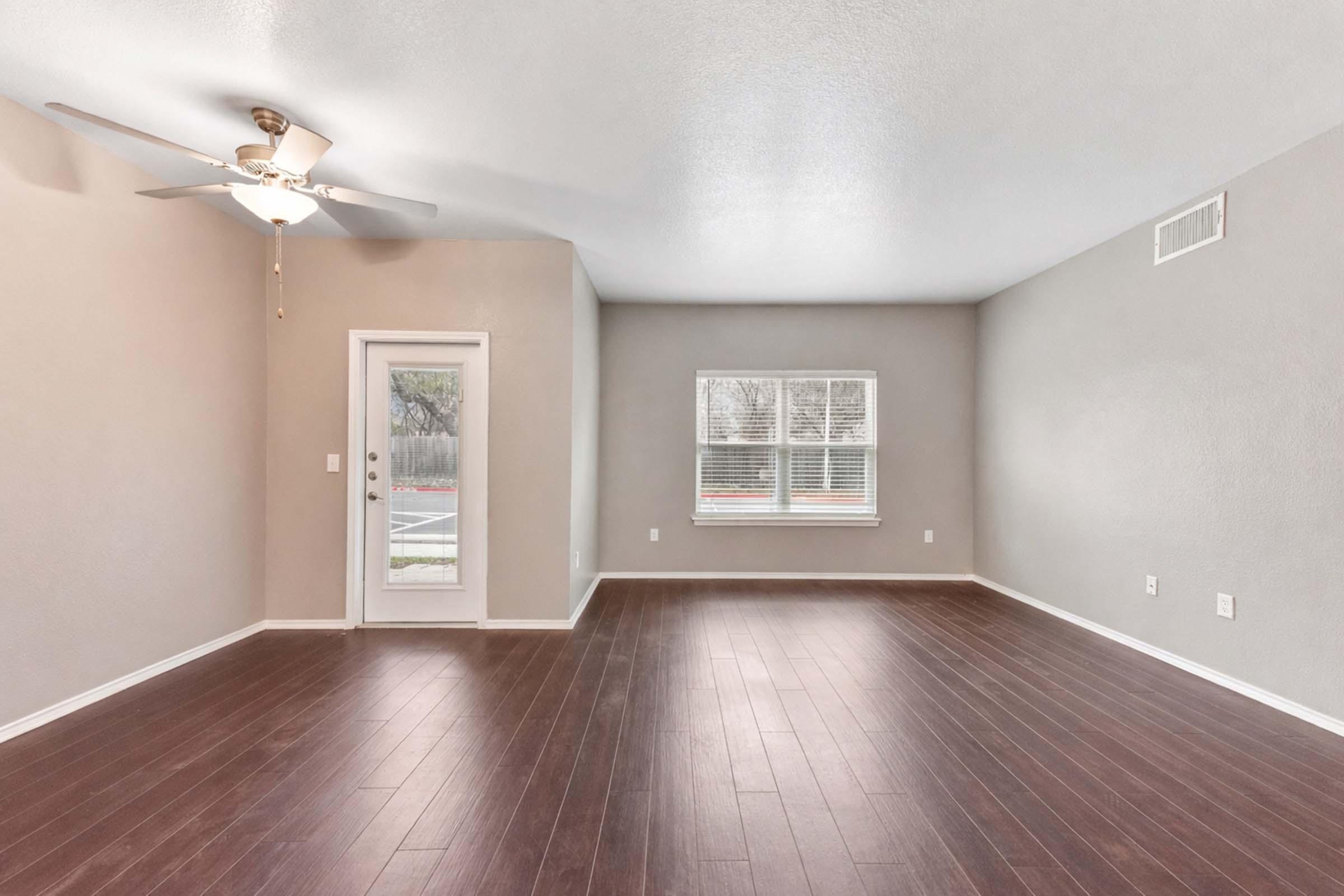 A spacious and empty living room featuring dark hardwood flooring, a ceiling fan, a single door leading outside, and a large window with blinds. The walls are painted in a light neutral color, creating a bright and airy atmosphere.