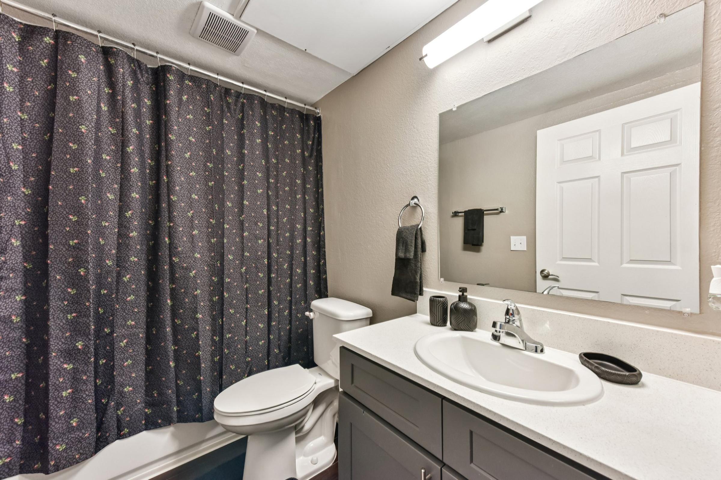 A clean and modern bathroom featuring a white sink with a dark cabinet, a toilet, and a mirror above the sink. The space is enhanced by a floral patterned shower curtain, a towel hanging on the wall, and minimal decor. Bright lighting illuminates the neutral walls, creating a welcoming atmosphere.