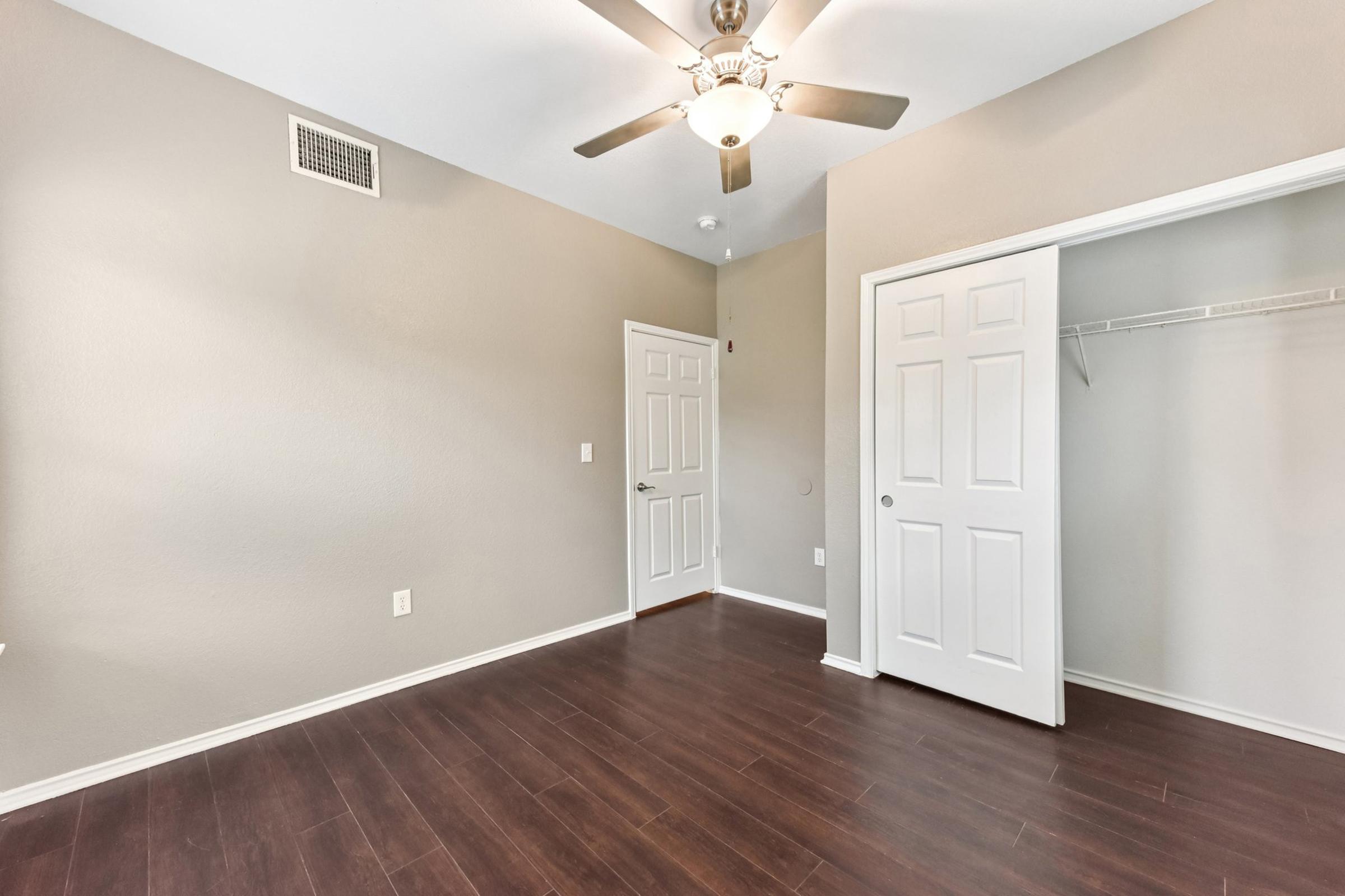 Empty room with light brown walls and a wooden floor. A ceiling fan with lights is installed above. There are two white doors: one leading to the closet and the other to the hallway. Natural light illuminates the space, highlighting the simplicity and neatness of the room.