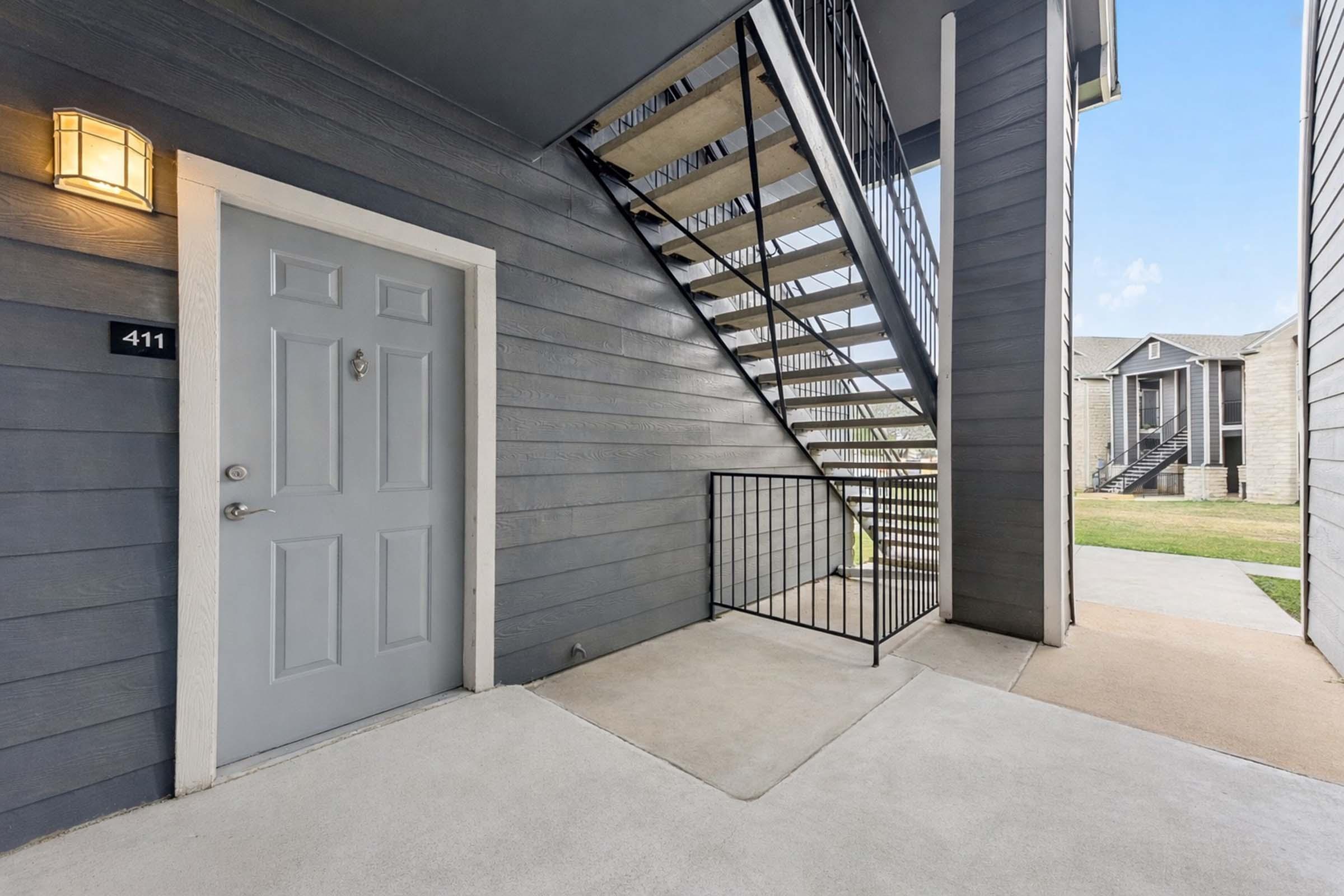 Entrance to an apartment featuring a gray door marked "411," a simple welcome mat, and a metal railing. A staircase leads to the upper level, with a view of grassy areas and other buildings in the background. The design is modern and clean, with a bright, clear sky above.