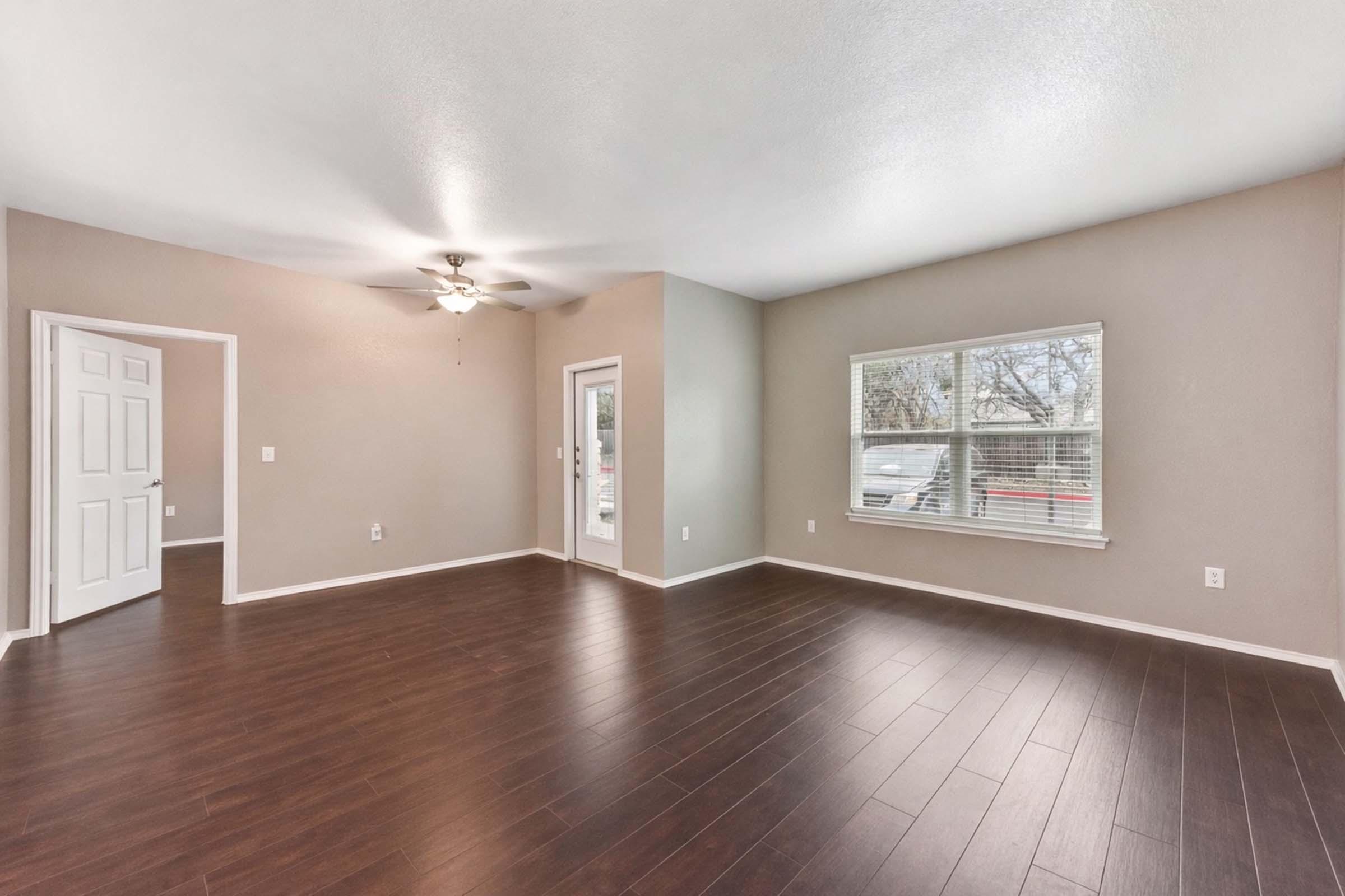 Spacious living room with hardwood floors, a ceiling fan, and neutral-colored walls. A large window allows natural light, while a door leads to an exterior entry. The layout includes an open space suitable for various furniture arrangements.