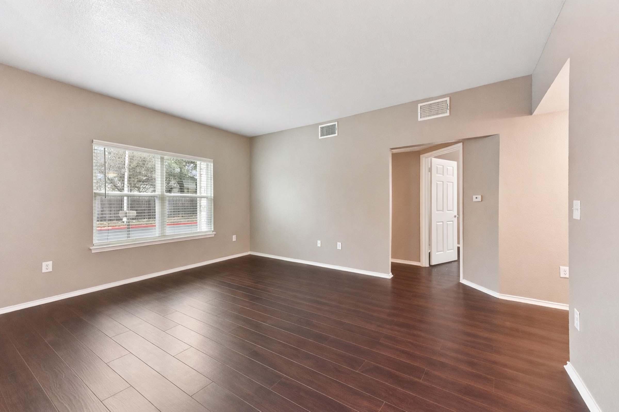 A spacious, empty room with light brown walls and dark wooden flooring. A large window allows natural light to enter, while white trim outlines the window and doorways. There is an open doorway leading to another area, creating a simple, airy atmosphere.