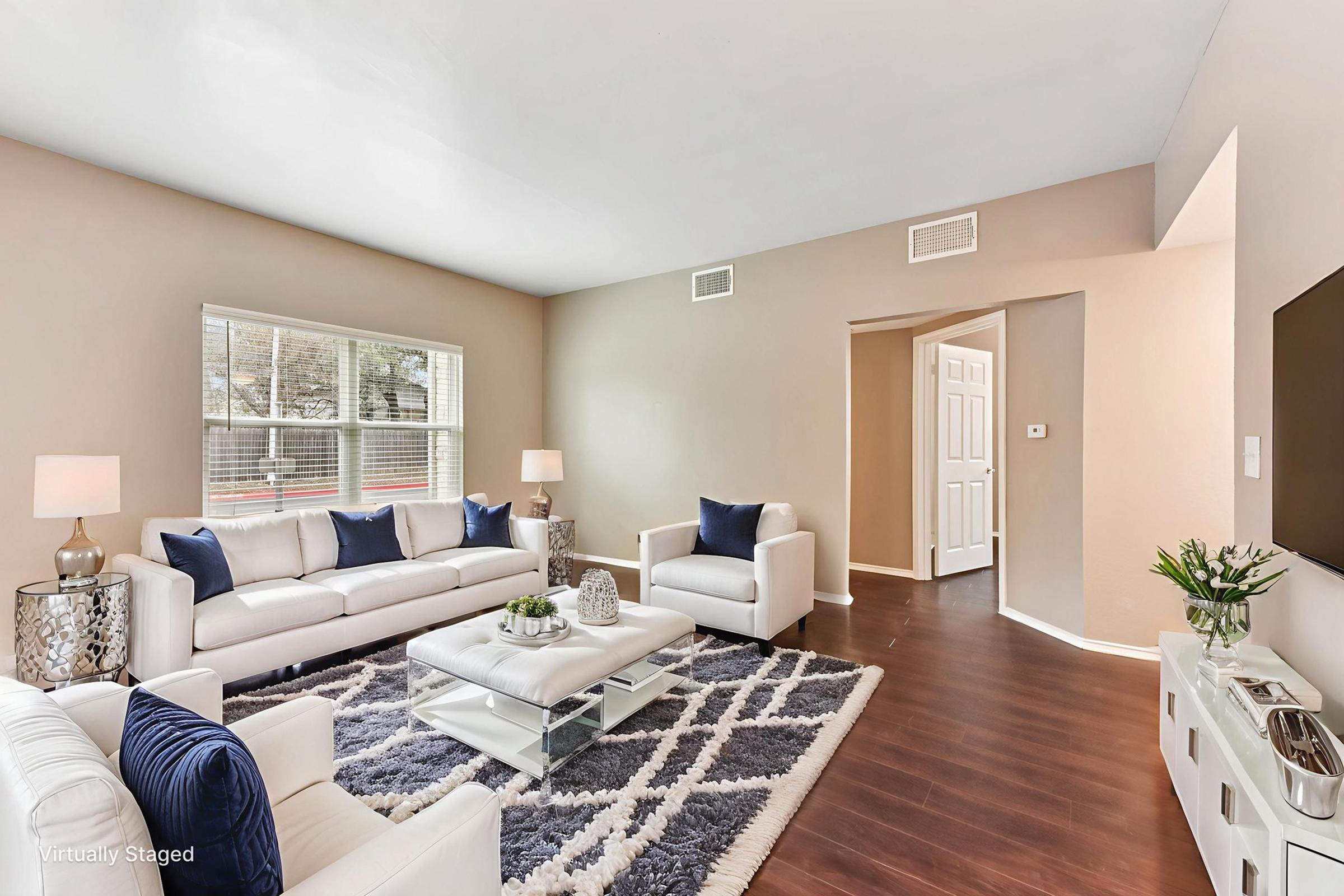 A stylish, modern living room featuring a light-colored sofa set with blue throw pillows, a plush area rug, a coffee table with decorative items, and lamps. The room has large windows allowing natural light, neutral walls, and an open layout that leads to another room.