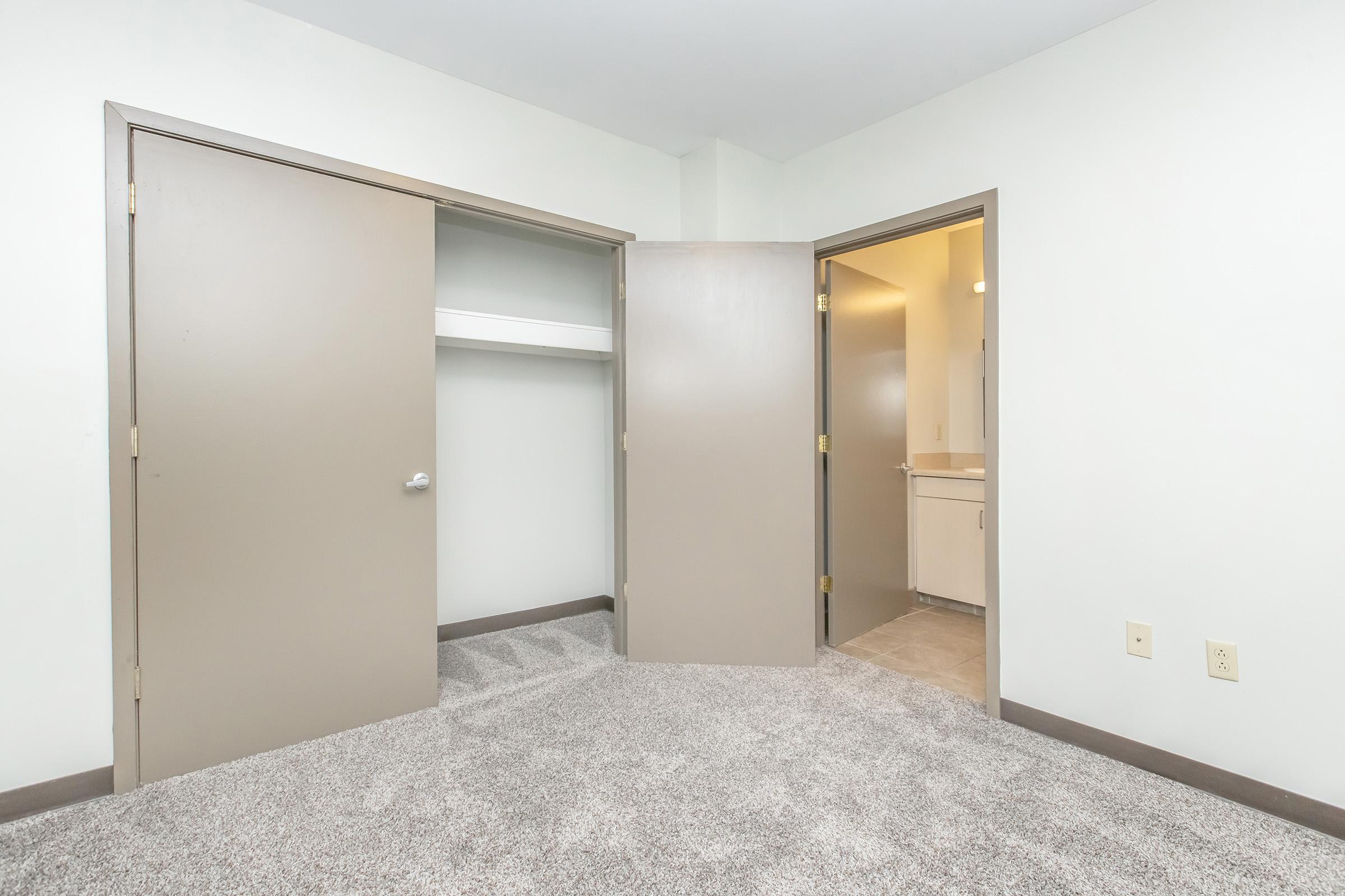A small interior room featuring two open doors. One door leads to a closet with a shelf, while the other opens to a bathroom area. The room has light-colored walls and a carpeted floor, creating a cozy atmosphere.
