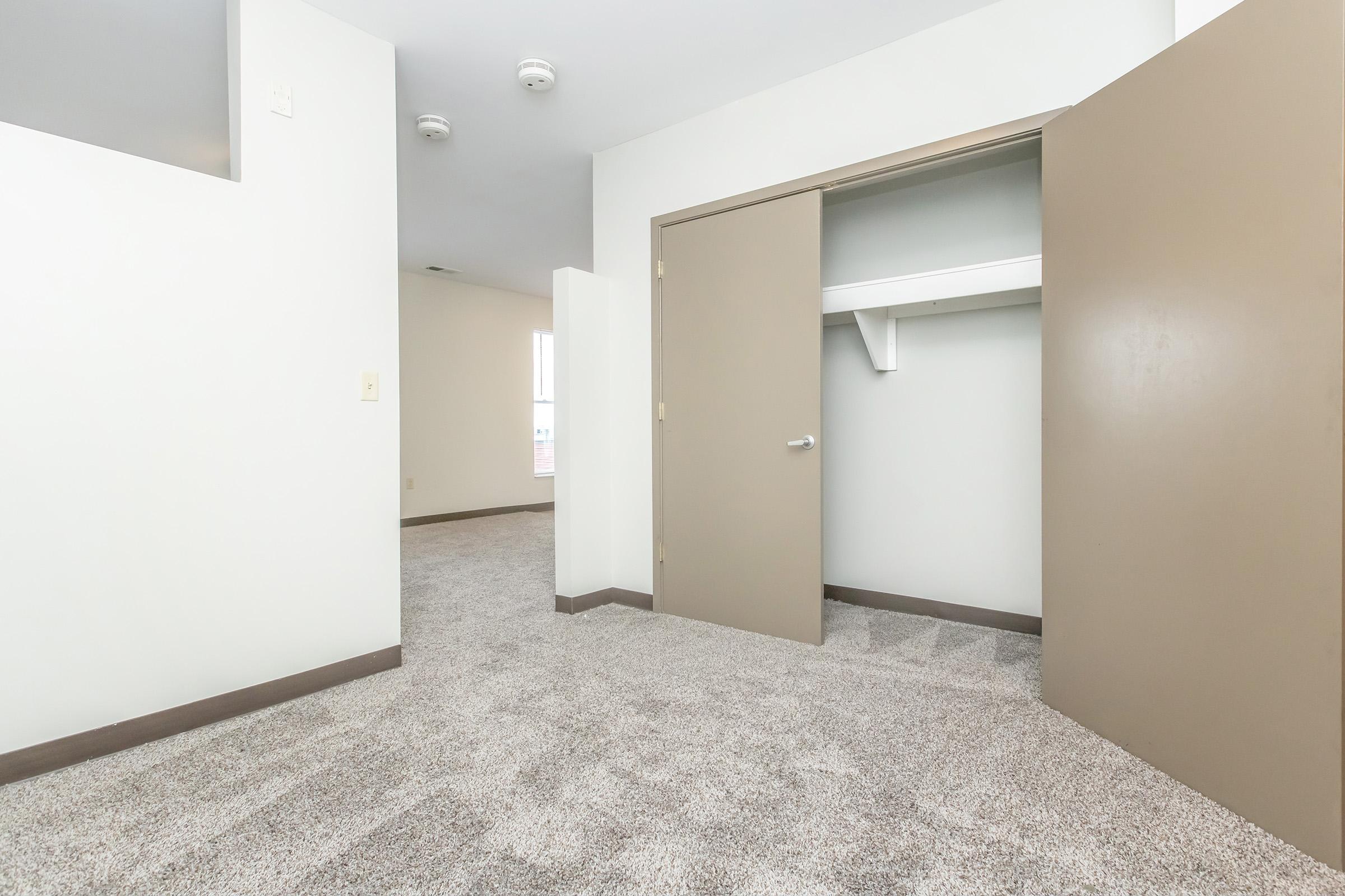 A well-lit room featuring a carpeted floor, a partially open closet with a shelf inside, and light-colored walls. The door to the closet is brown, and the room has a simple, minimalist design with no furniture visible.
