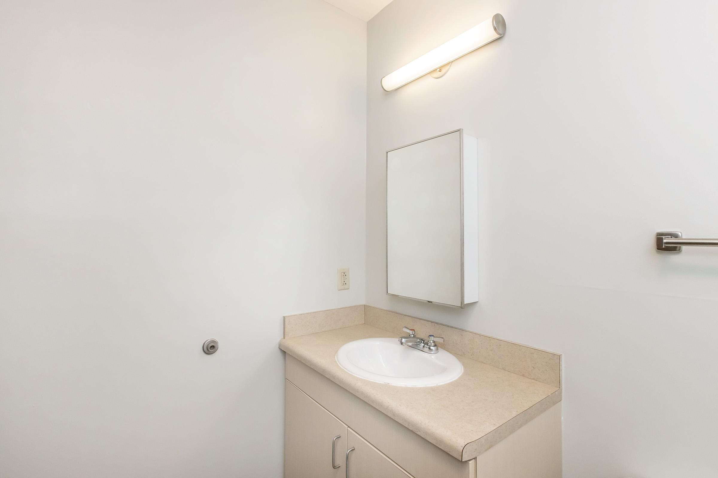 A simple bathroom with a light-colored countertop, a circular sink, and a rectangular mirror mounted above it. The walls are plain and light-colored, and there is a towel bar on the right. The overall atmosphere is clean and minimalistic, providing a functional space.