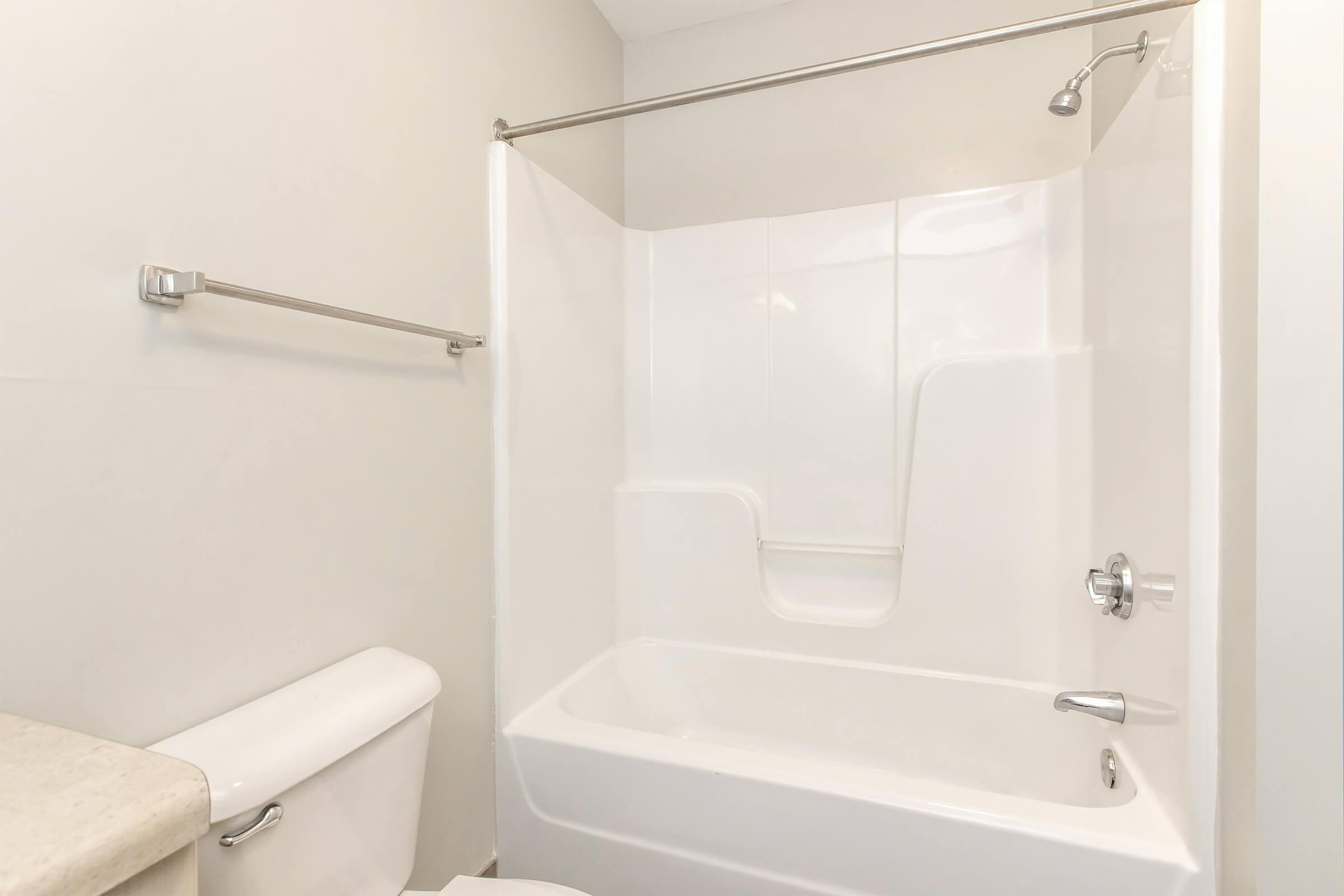 A clean, white bathroom featuring a bathtub with a showerhead above it, a chrome shower rod, and a toilet nearby. The walls are painted light gray, and the flooring is not visible. The space appears modern and well-maintained.
