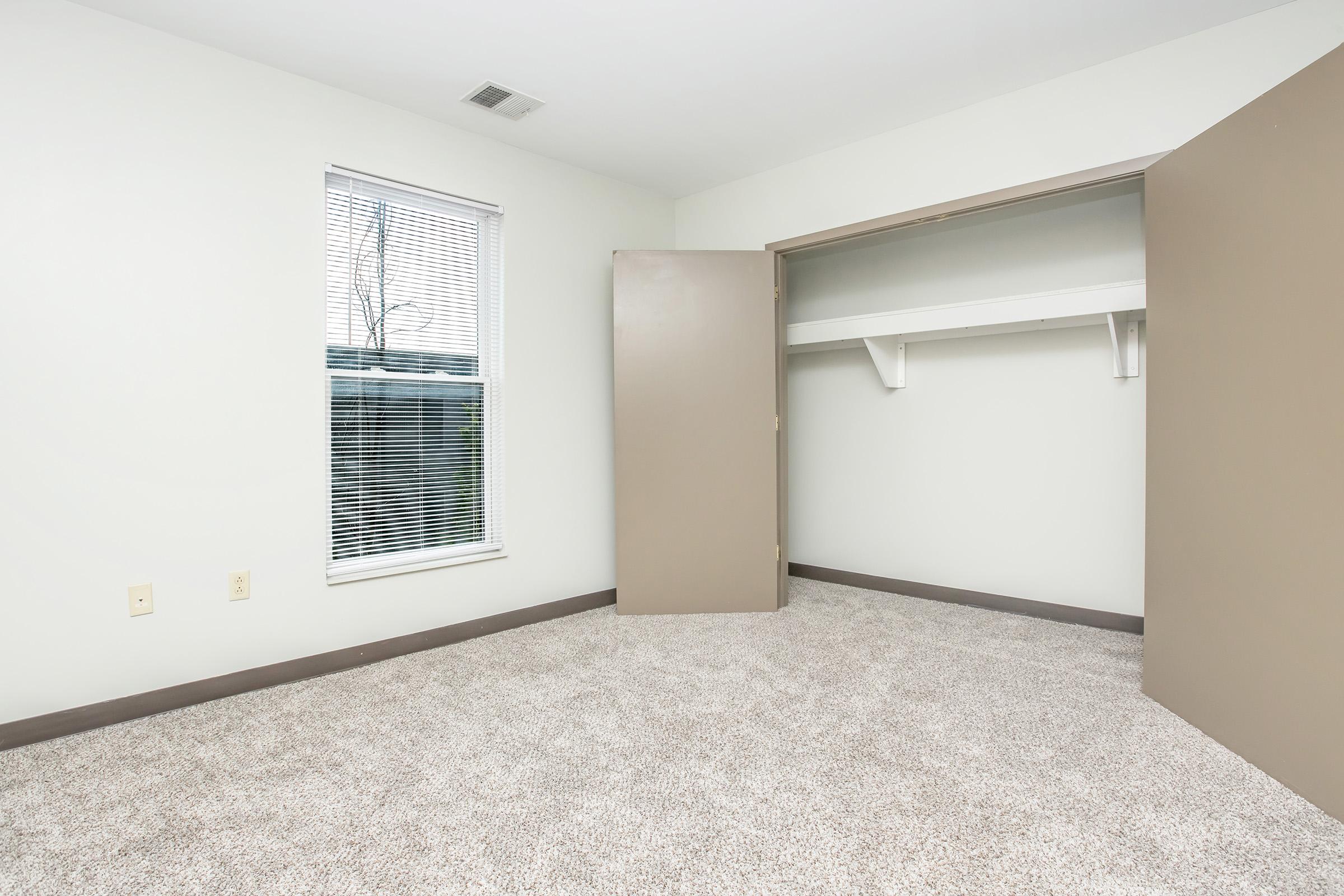 A spacious, empty room featuring light-colored walls and carpet. There's a window with blinds letting in natural light, and an open closet with two doors, displaying a shelf inside. The space is well-lit and clean, ideal for personal customization.