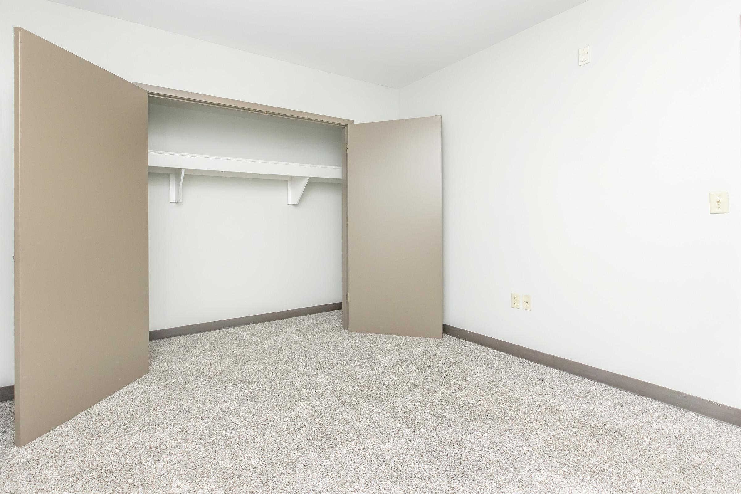 A spacious, empty room featuring beige walls and carpet. An open closet with two doors reveals a shelf and a clean, uncluttered floor area, creating a minimalist aesthetic. Natural light brightens the space, highlighting its simplicity and potential for personal customization.