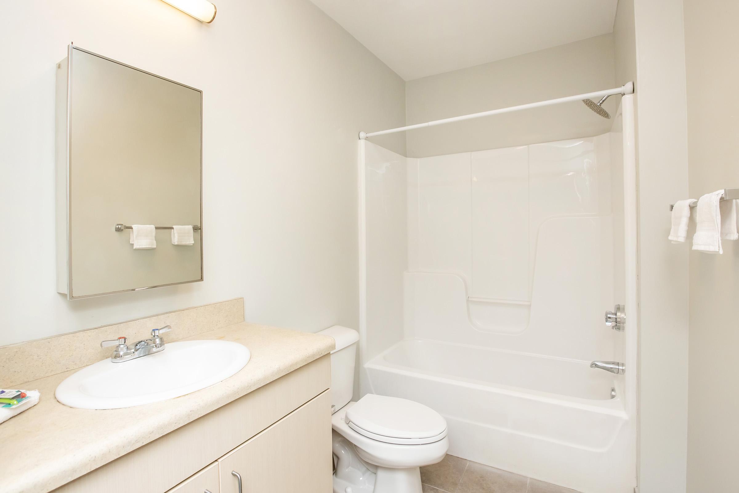 A clean and modern bathroom featuring a white bathtub with a shower, a sink with a countertop and a mirror above it. The walls are painted in a light color, and there are neatly arranged towels hanging on a towel rack. The overall ambiance is bright and minimalist.