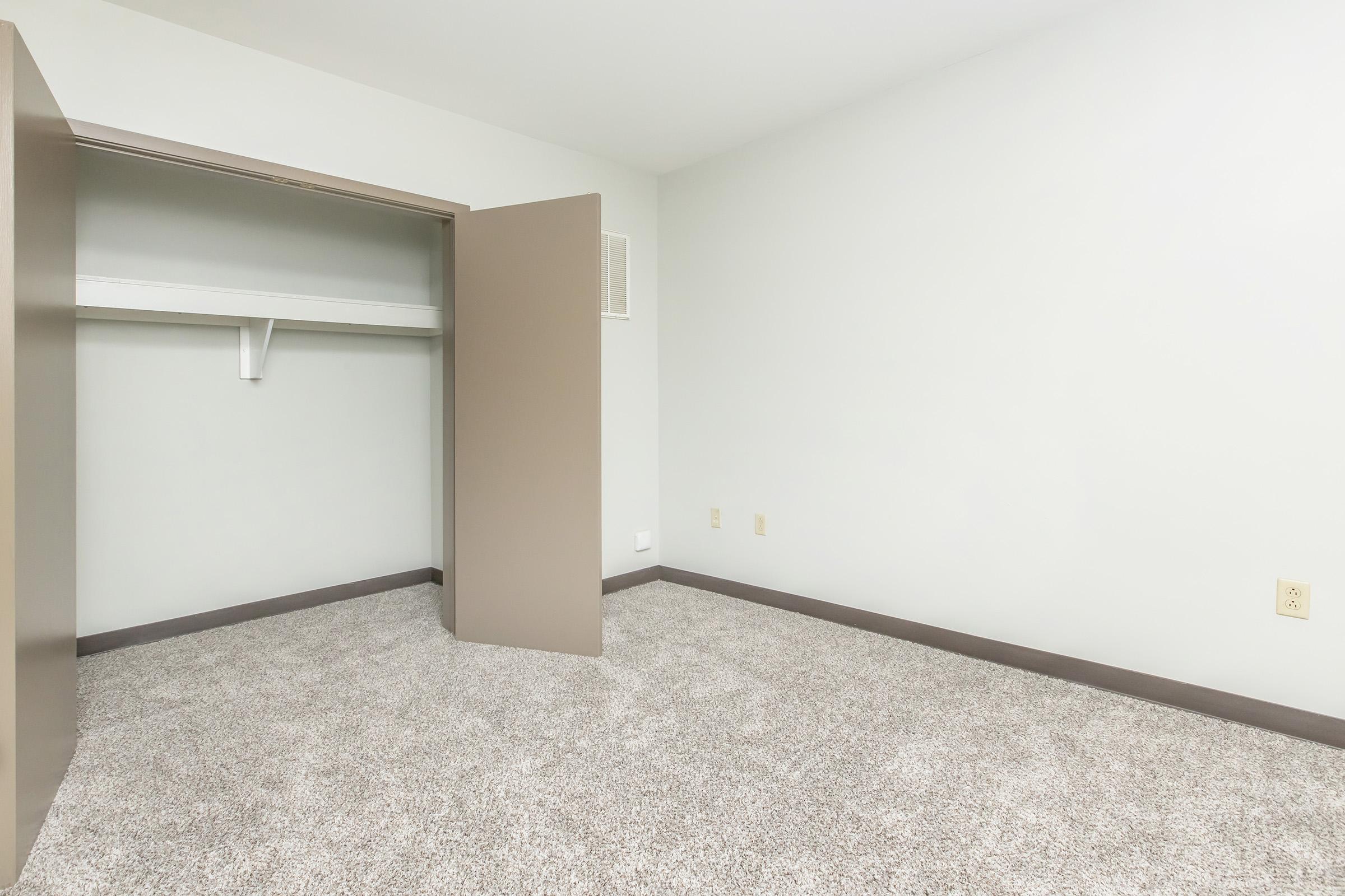 Empty room with light gray walls and beige carpet. A closet with open double doors is on the left, featuring a shelf inside. The room has no furniture, creating a spacious and minimalist atmosphere.
