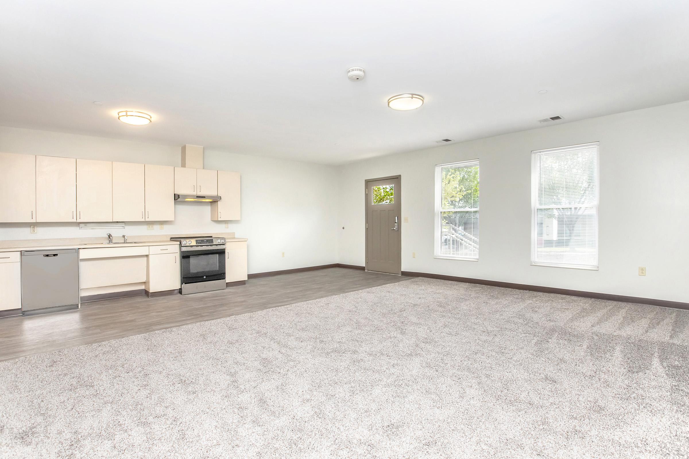 A spacious, well-lit room featuring a kitchen area with light-colored cabinets, a stove, a dishwasher, and a door leading outside. The flooring is carpeted, and the walls are painted light colors, creating a clean and inviting atmosphere with large windows allowing natural light.