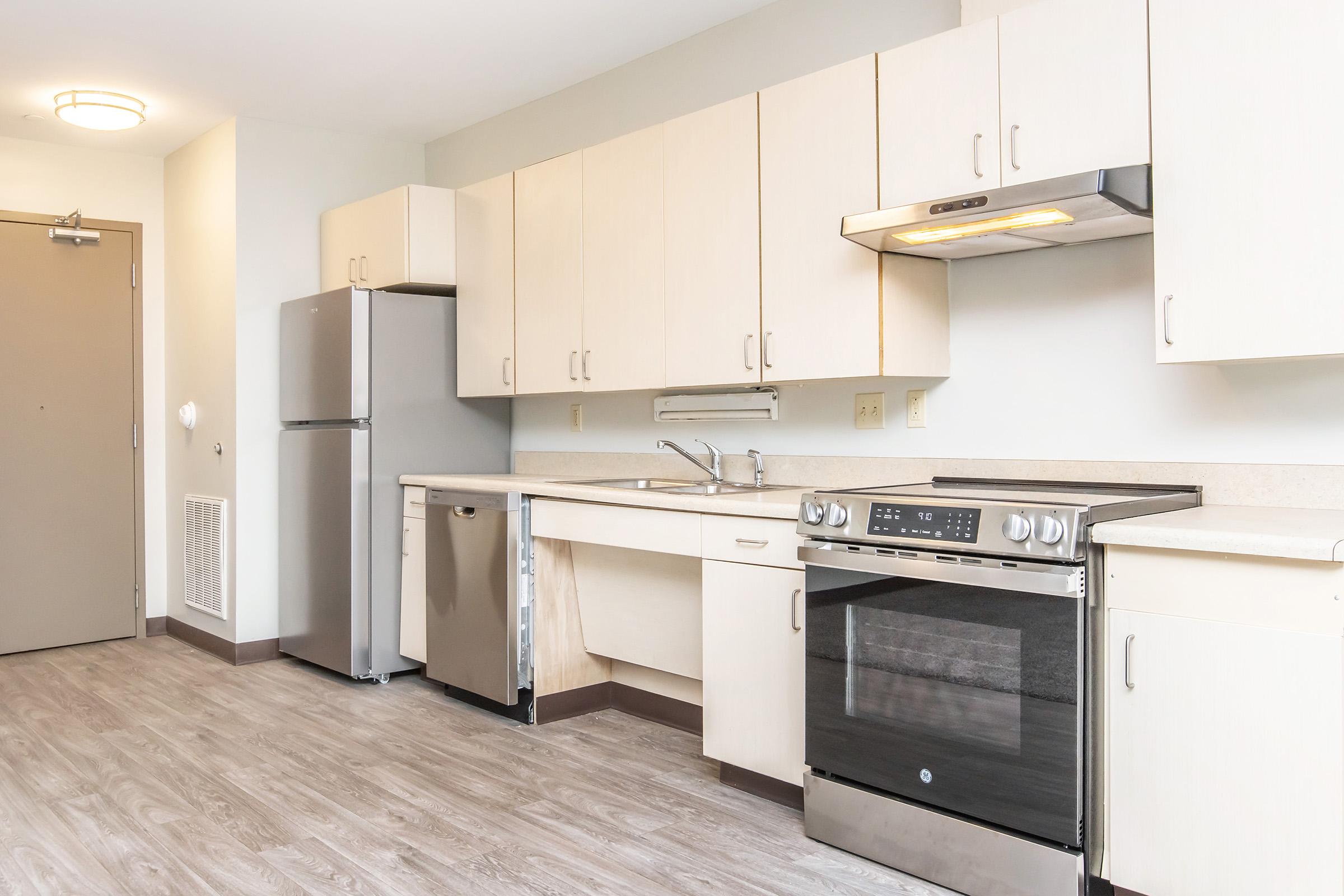 A modern kitchen featuring light-colored cabinets, a stainless steel refrigerator, a dishwasher, and a stovetop with an oven. The countertops are neutral, and the flooring is a wood-like laminate. There is a door leading outside and a ceiling light fixture visible.