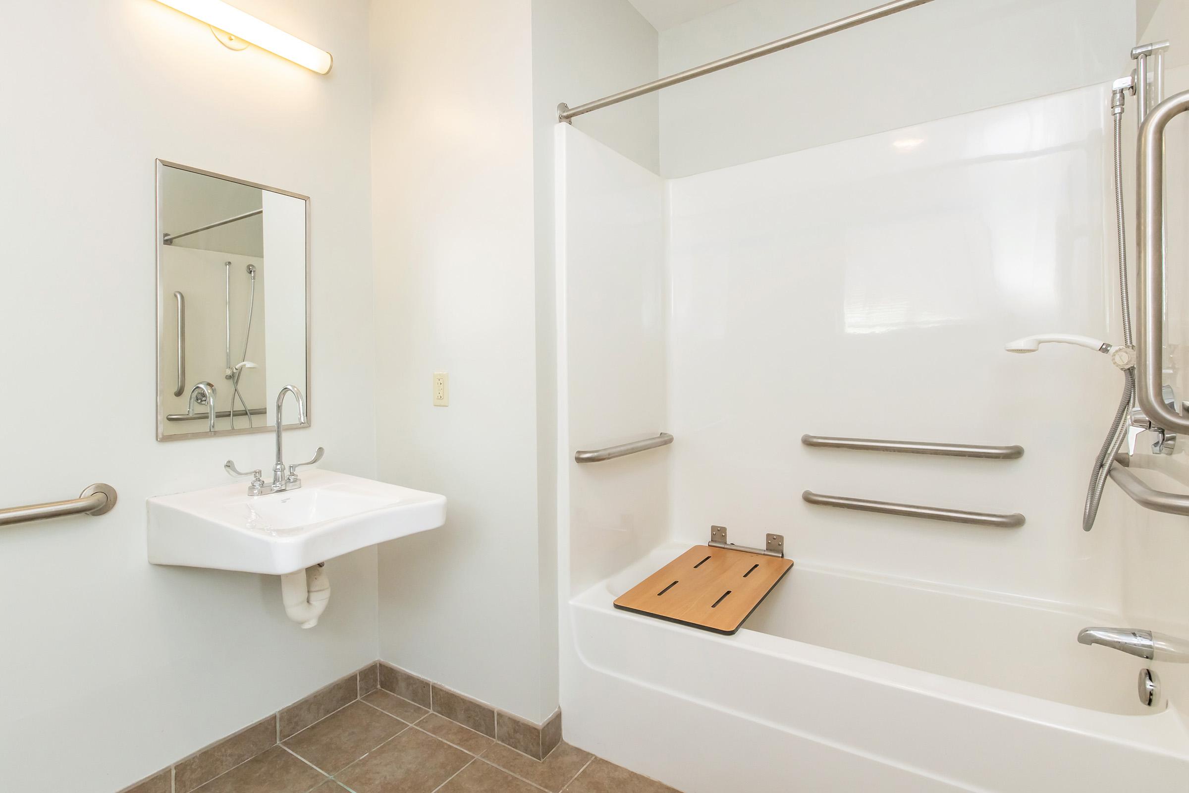 A clean, modern bathroom featuring a bathtub with a wooden transfer bench, accessible grab bars, and a small sink. There is a mirror above the sink, and the walls are painted light colors, creating a bright and spacious atmosphere. The flooring is tiled in neutral tones.