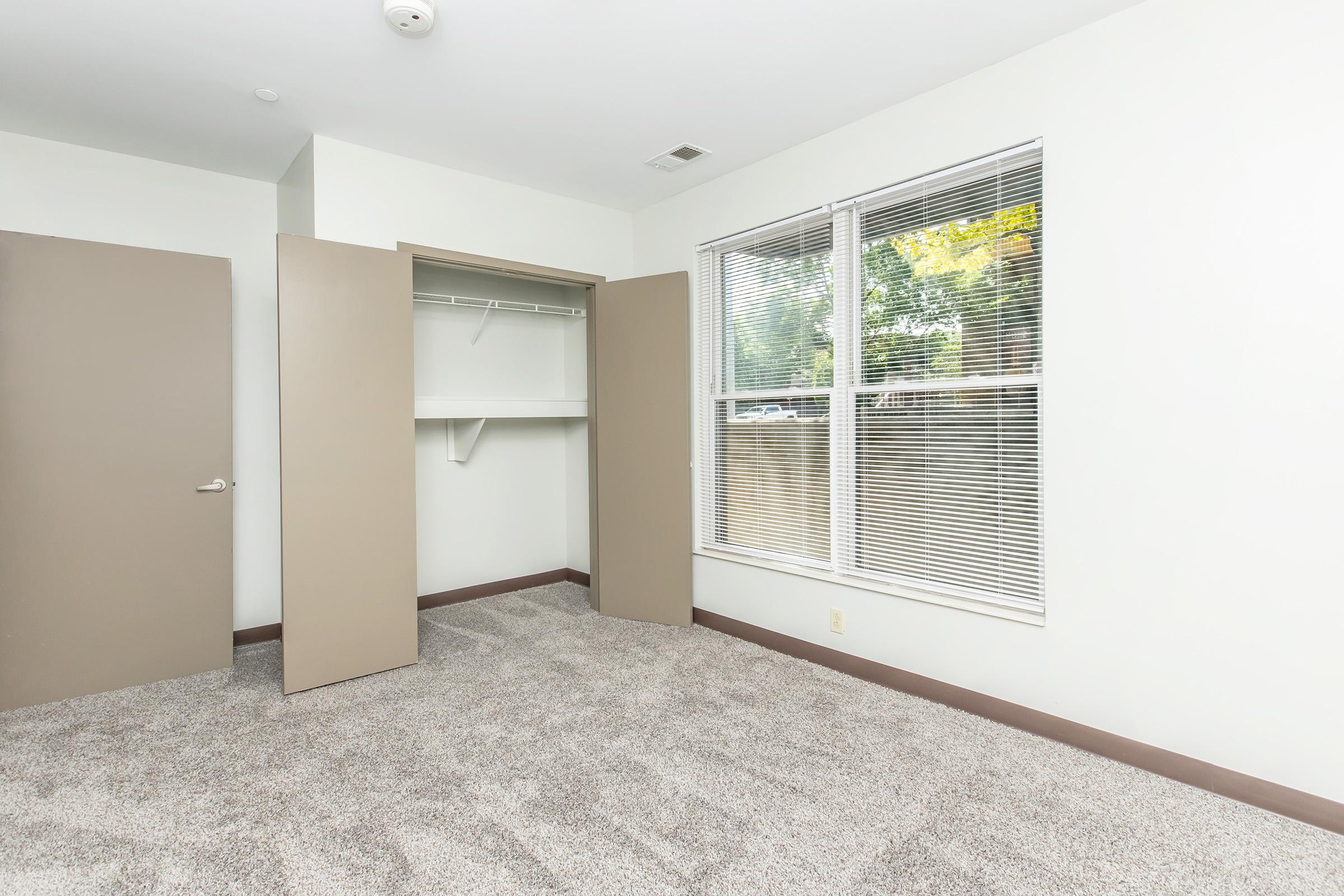 Empty room featuring gray carpet, white walls, and a large window with blinds. A two-door closet is visible on the left side, and the room has a clean, minimalist look with natural light coming in.
