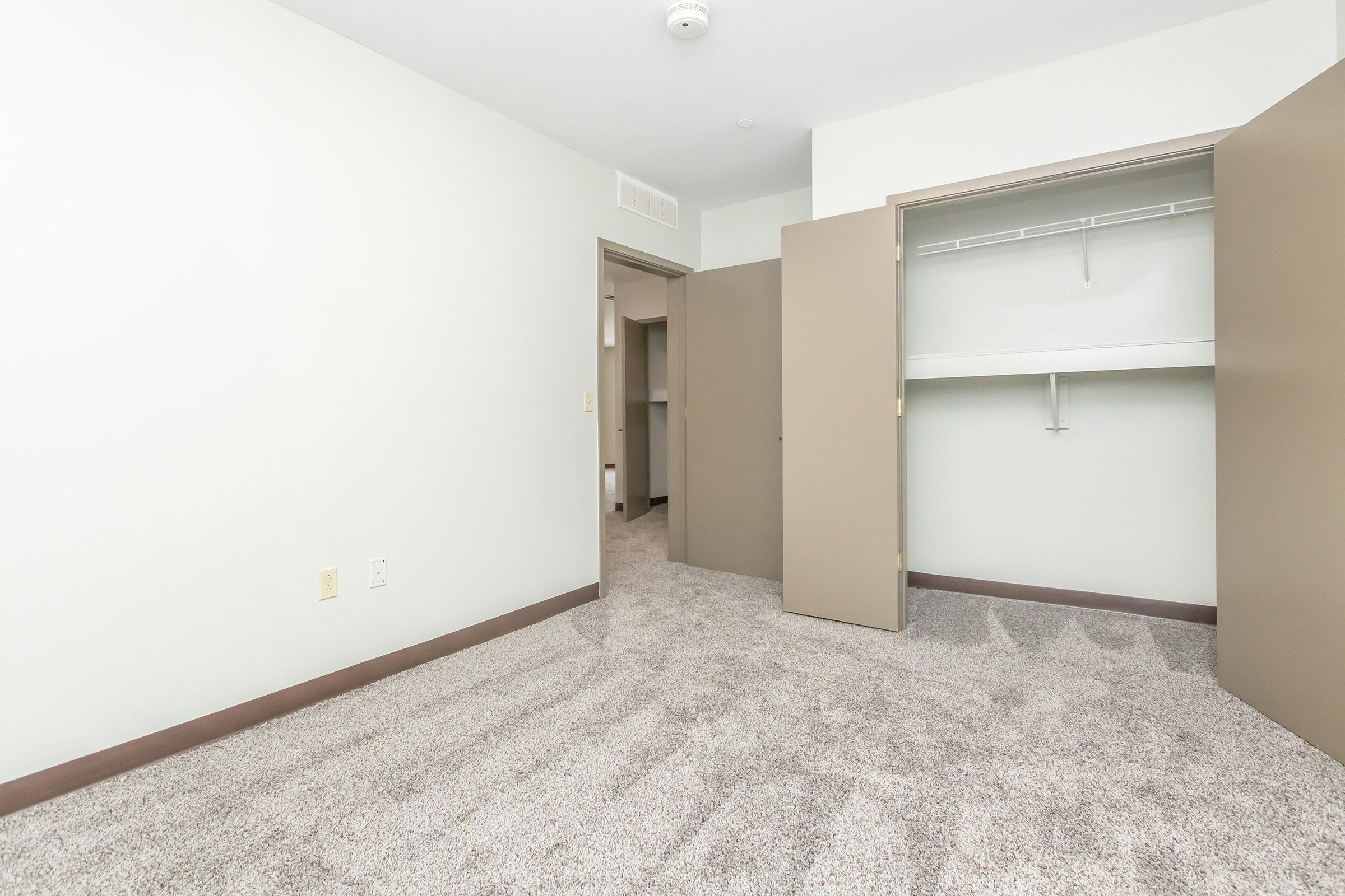Empty room with light-colored walls and carpeted flooring. There are two open doors leading to a closet with a shelf. The space is well-lit and features a neutral color scheme, suggesting a clean and modern design.