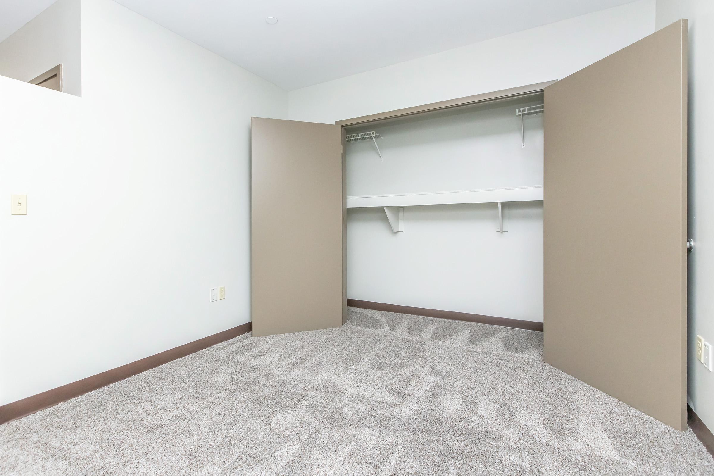 An empty closet with two open doors, featuring a hanging rod and a shelf above, set against a light-colored wall. The floor is covered with plush, neutral-colored carpet.