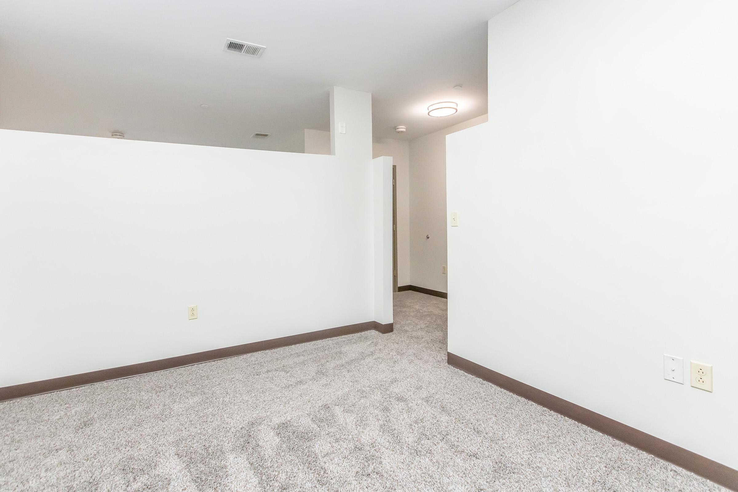 A spacious, unfurnished room with light gray carpet and white walls. There are partially visible walls creating a sense of separation, a ceiling light fixture, and an entrance to another area. The overall ambiance is clean and modern, suitable for various interior designs.