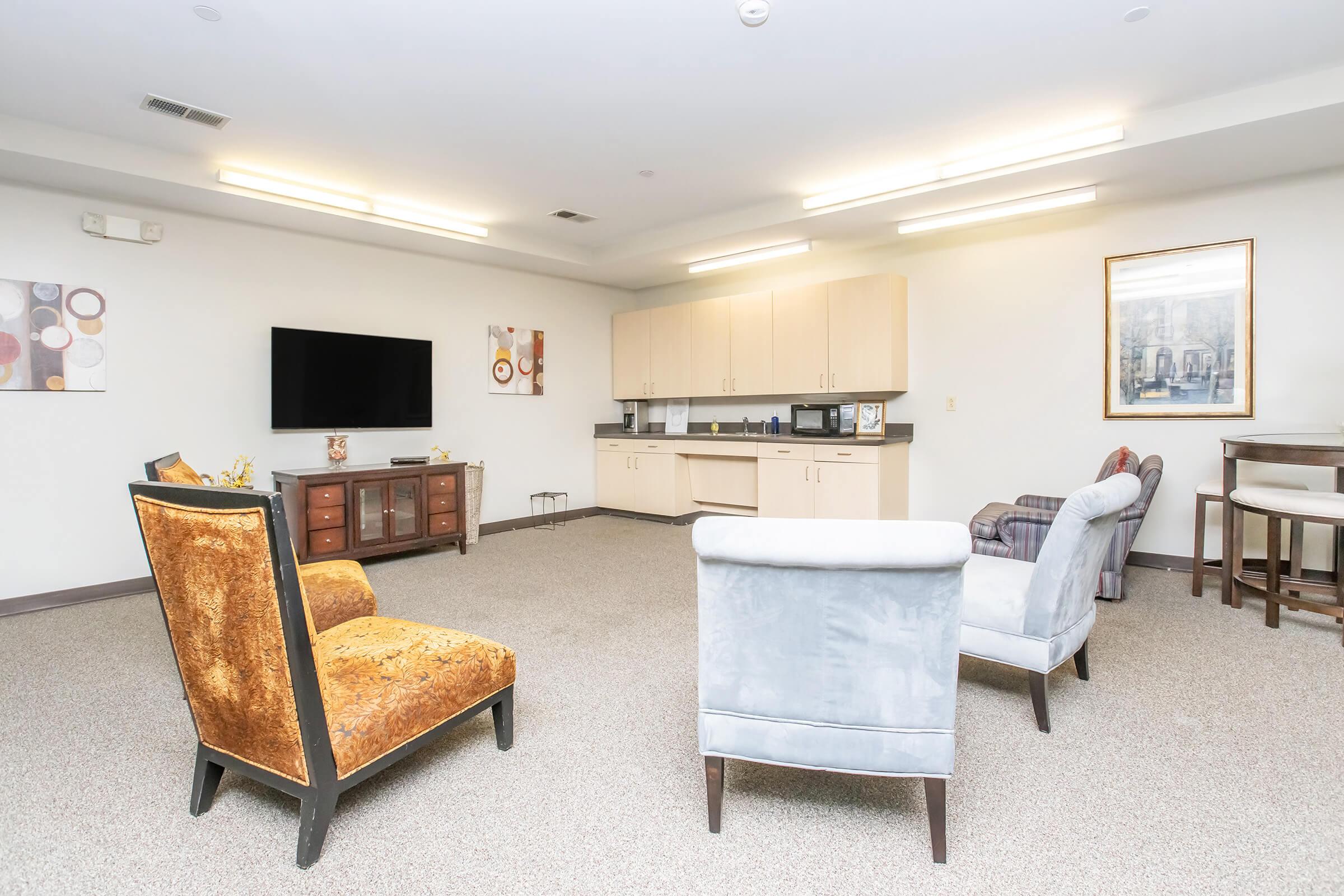 A spacious room featuring four upholstered chairs arranged in a circle, a wooden cabinet, a wall-mounted TV, and a kitchenette area with light-colored cabinets. Neutral walls and carpet enhance the bright ambiance. A framed artwork is visible on one wall.
