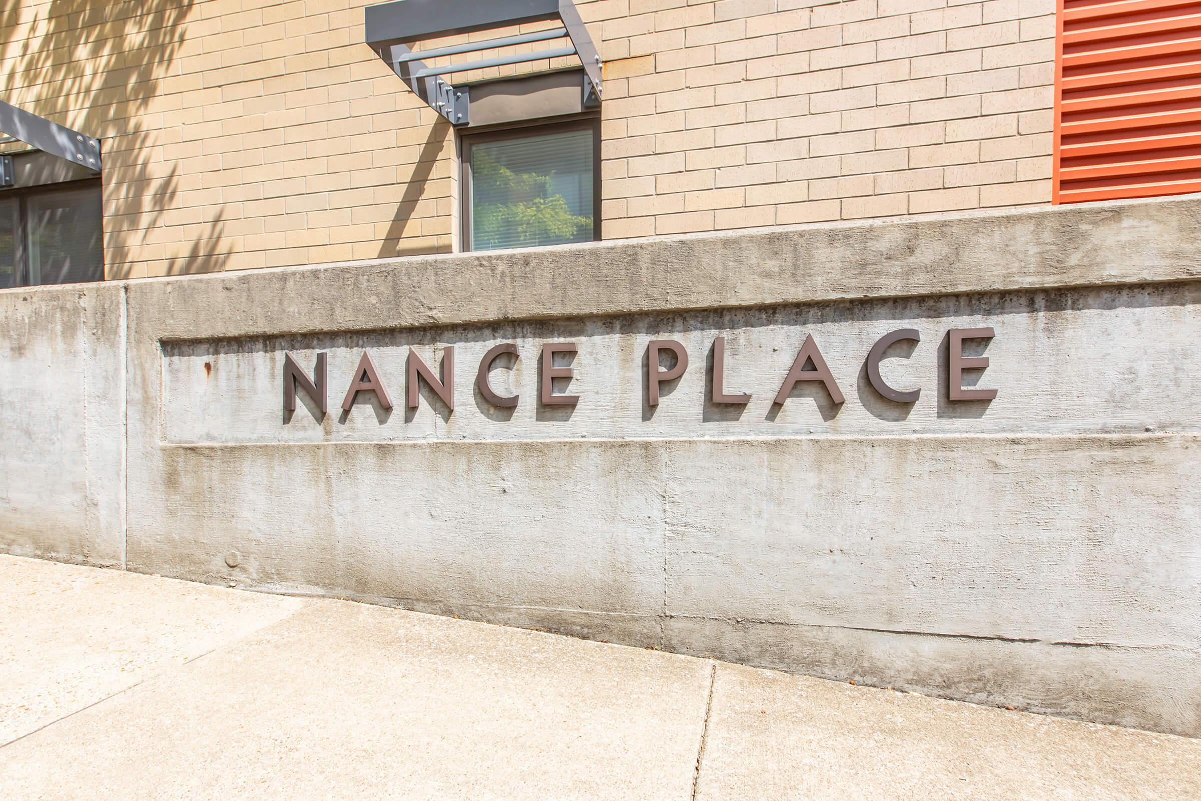 Concrete wall with the engraved text "NANCE PLACE" in bold letters, situated near a modern building with a light-colored exterior. Natural light casts soft shadows on the wall, and there are hints of greenery in the background.
