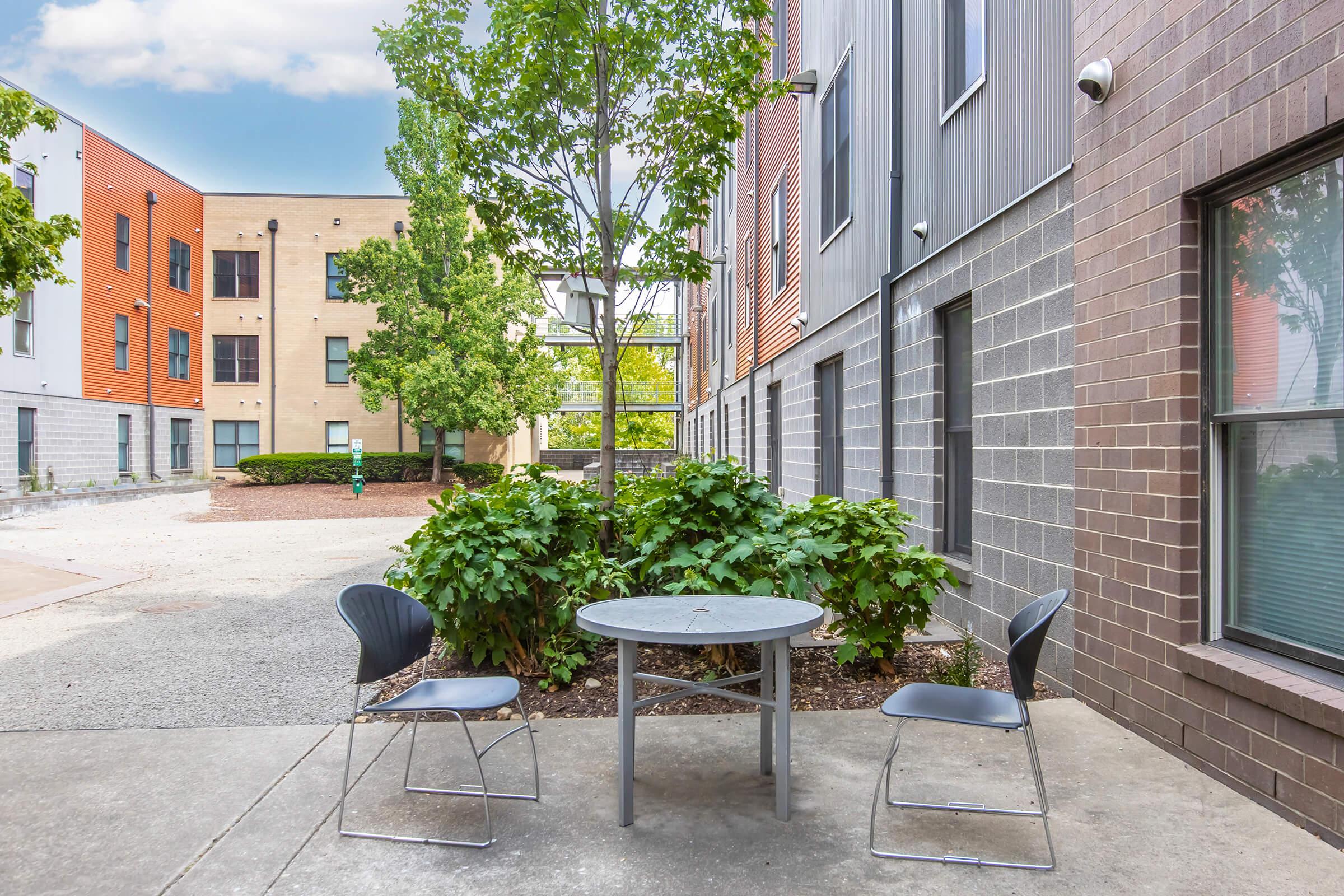 A small outdoor seating area featuring a round table and two metal chairs surrounded by greenery. In the background, there are modern buildings with a mix of brick and colorful facades. The scene is bright and inviting, with clear blue skies and a hint of greenery from nearby trees.