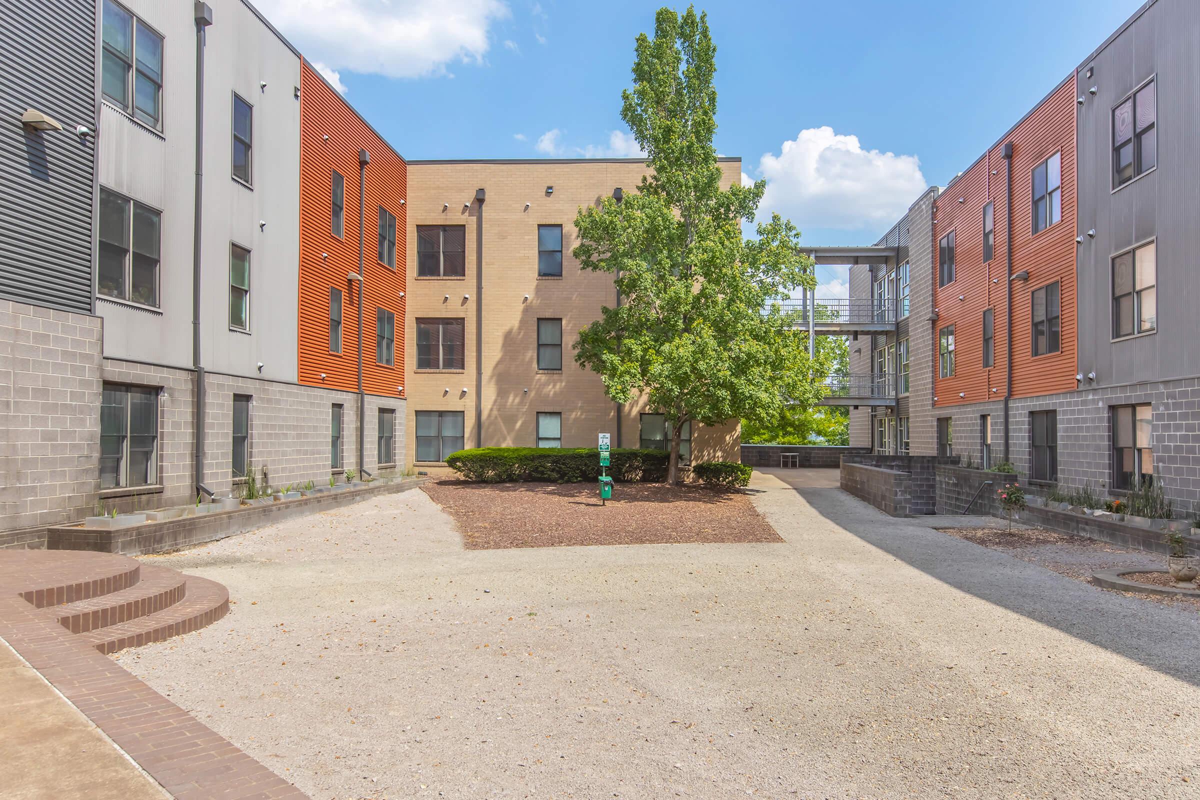 A view of a modern multi-story residential complex with a courtyard. The courtyard features gravel and landscaping, surrounded by buildings with a mix of brick and colorful siding. Trees are present, and blue skies and clouds are visible in the background.