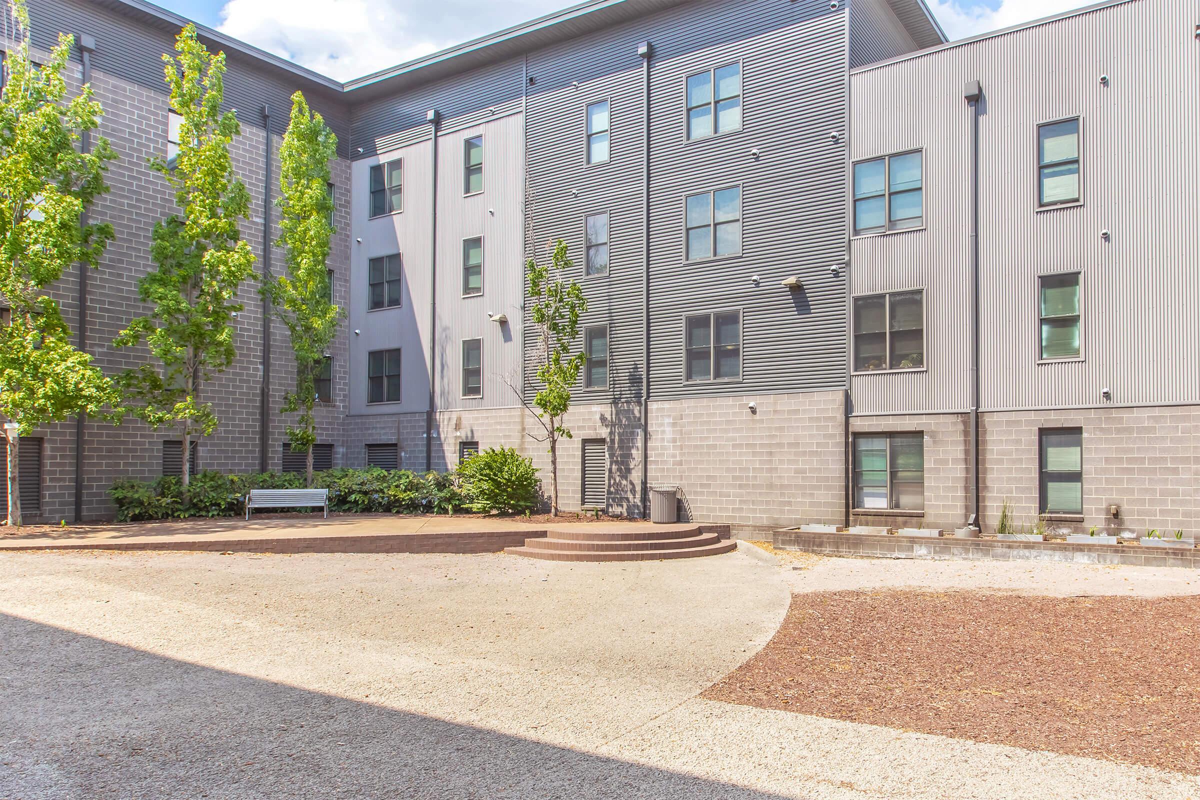 A modern, multi-story building with a gray exterior featuring several windows. In the foreground, there is a landscaped area with gravel and a few small trees, alongside a set of circular steps leading to an entrance. Sunshine illuminates the courtyard, creating a bright and open atmosphere.
