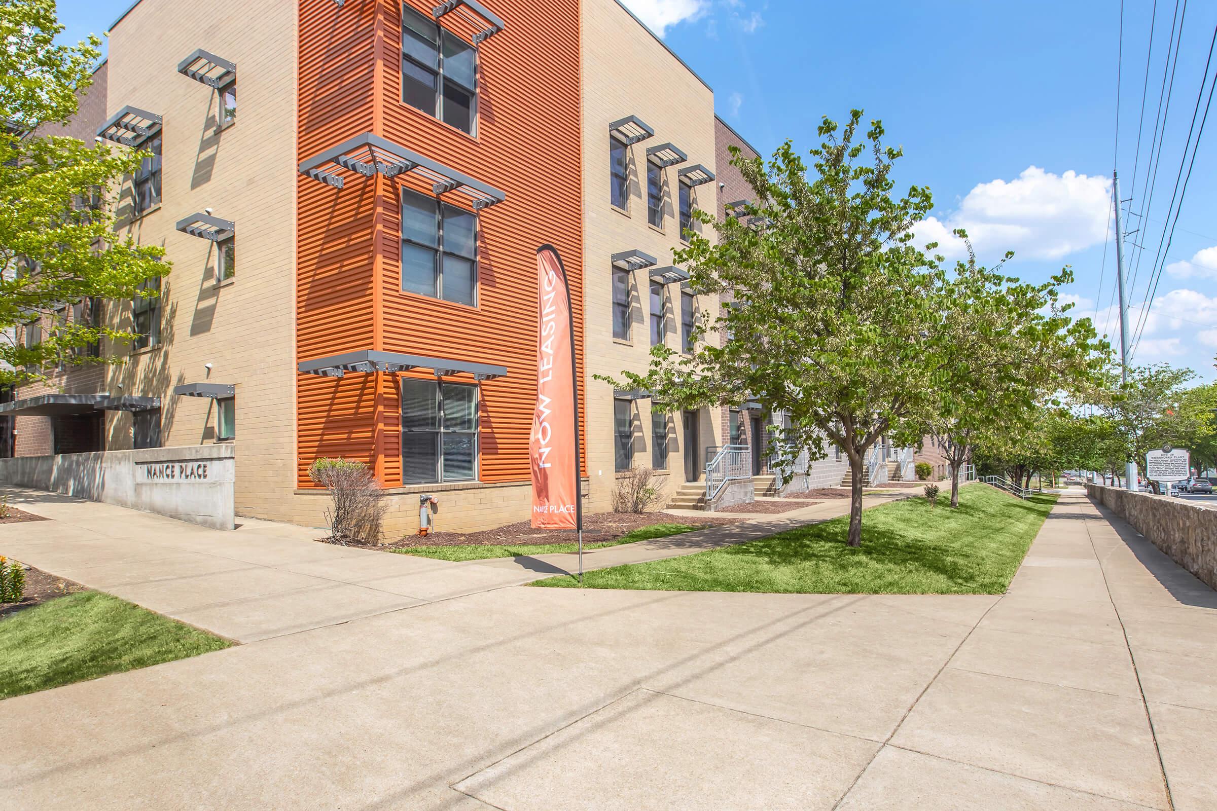 A modern building with a combination of orange and beige exterior, featuring large windows and a banner. Surrounding the structure are neatly maintained sidewalks, grassy areas, and trees. The scene is bright and sunny, highlighting the architecture and landscaping.
