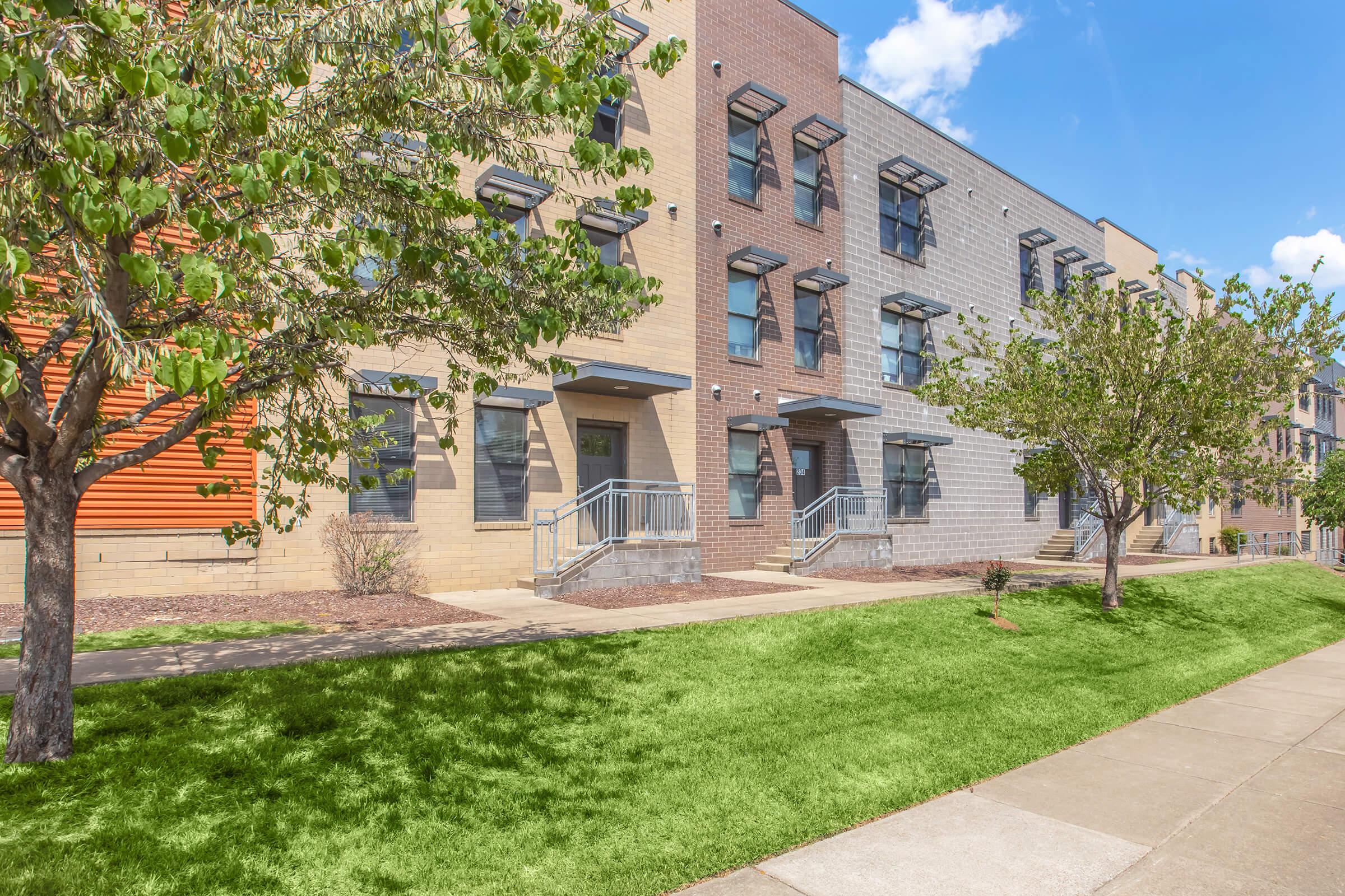 A modern, two-story building featuring a mix of brick and gray siding. Large windows and balconies are visible. The area is landscaped with grass and small trees, alongside a concrete pathway. The sky is bright with a few fluffy clouds.
