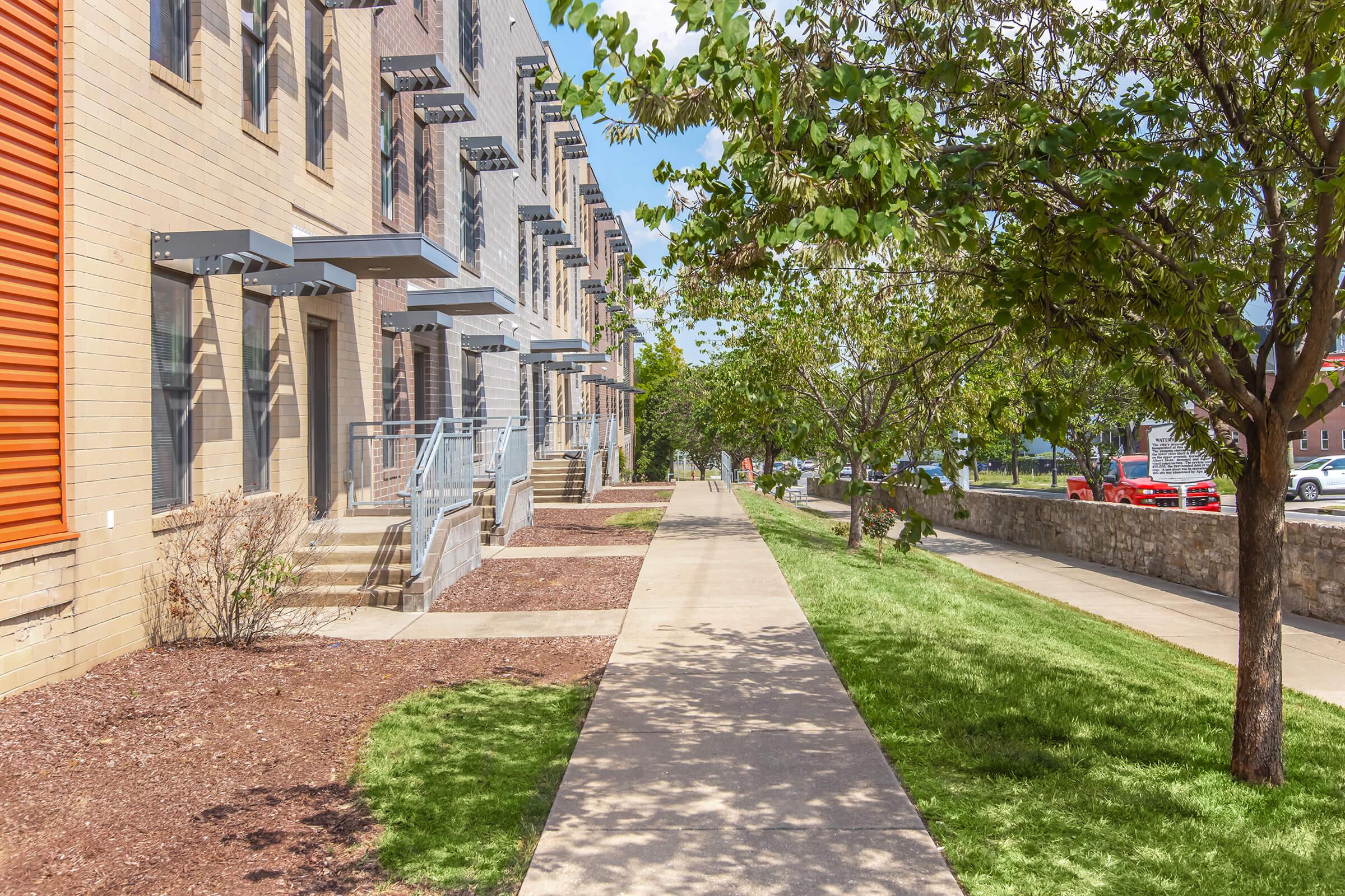 A paved pathway lined with green grass and trees, leading past modern residential buildings with balconies. The scene is bright and sunny, showing a well-maintained neighborhood with a sidewalk on one side and a street visible on the other.