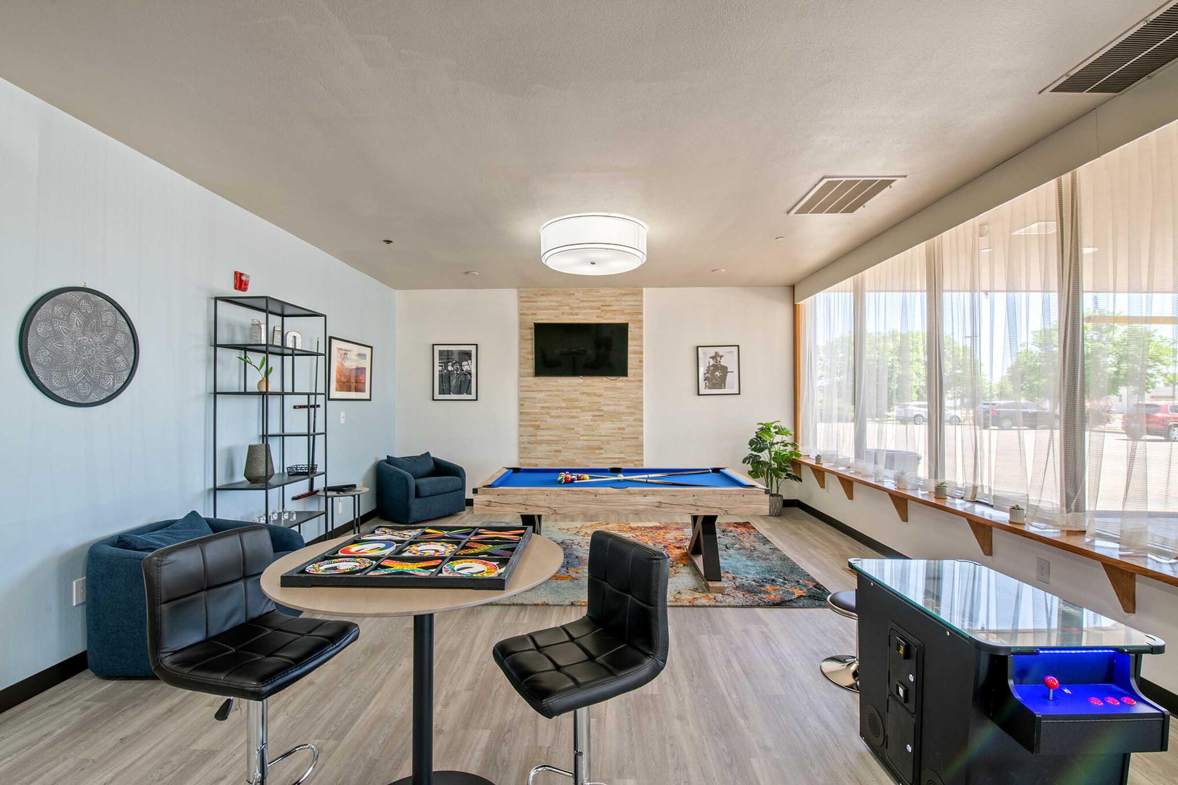A modern game room featuring a blue pool table, stylish seating, and a small dining area with bar stools. A decorative wall with art and a TV is visible, along with a console game machine. Large windows allow natural light to brighten the space, showcasing a relaxed social atmosphere.