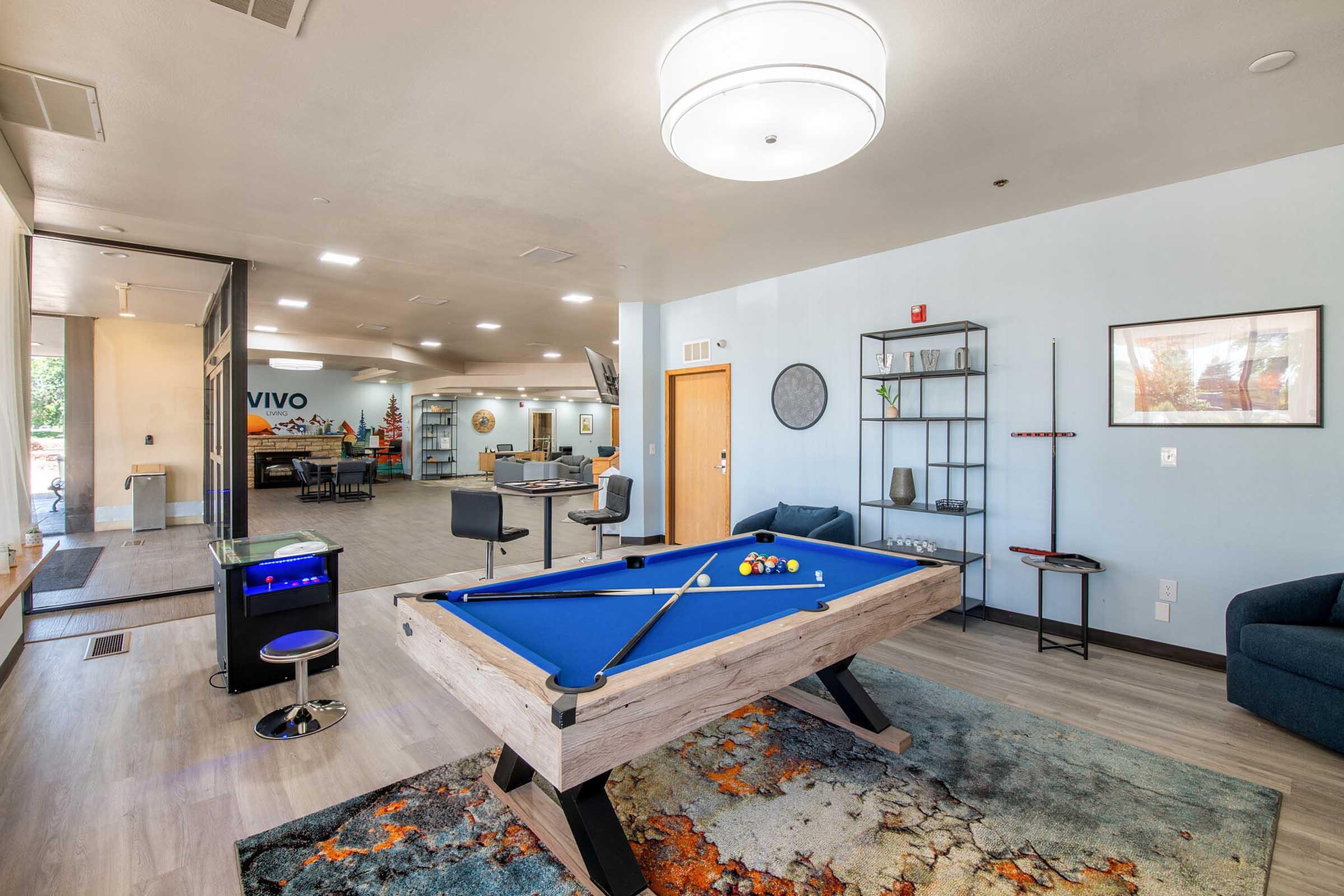 A modern lounge area featuring a blue pool table in the foreground, surrounded by a stylish rug. In the background, there are seating areas and a desk, with a large window allowing natural light. The walls are painted in soft colors, and decorative shelves add a contemporary touch.
