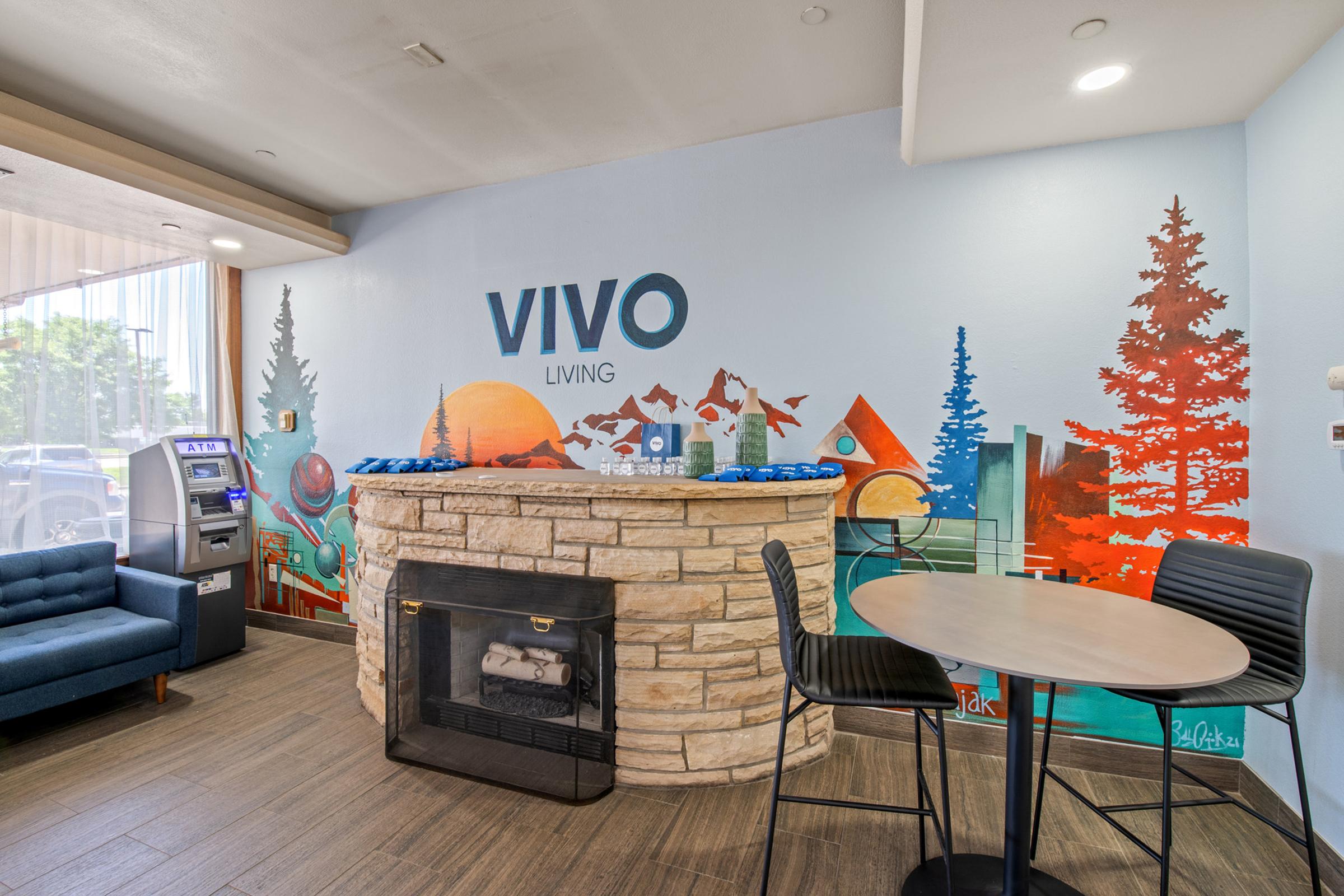 Interior scene of a lobby featuring a vibrant mural with mountains, trees, and abstract shapes. A stone fireplace is situated against the wall with a circular table and chairs nearby. A blue sofa is in the corner, and there's an ATM machine visible. The overall atmosphere is welcoming and modern.