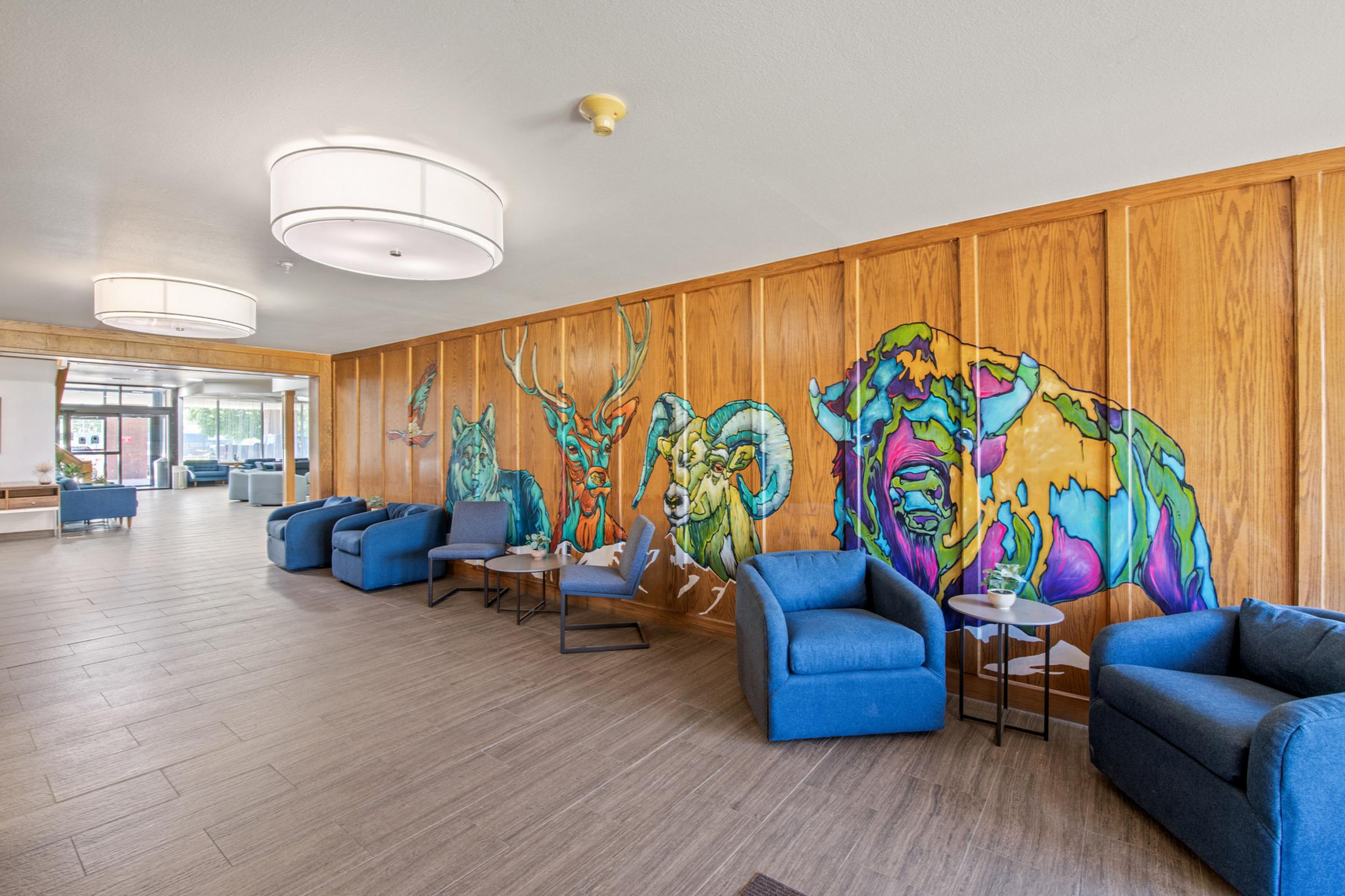A modern interior space with wooden paneling featuring vibrant animal murals, including a cat, deer, bison, and an abstract figure. The room has light blue chairs and small tables arranged for socializing, with large round light fixtures providing bright illumination. Natural light enters from windows in the background.