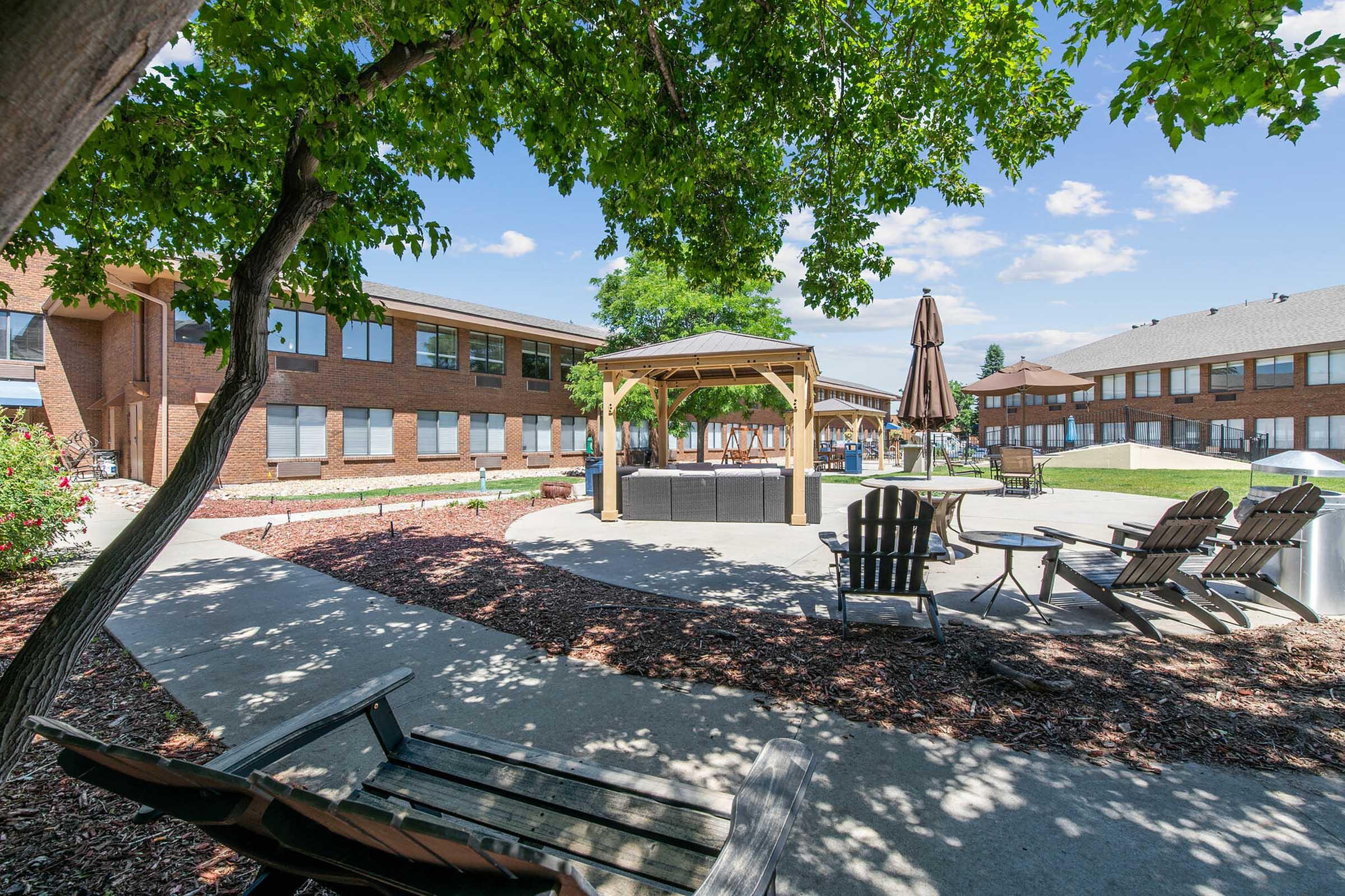 A serene outdoor space featuring a gazebo at the center, surrounded by well-maintained landscaping and chairs. The scene includes a few trees providing shade, with a sunny sky and clouds in the background, creating a relaxing atmosphere for gatherings or leisure activities.