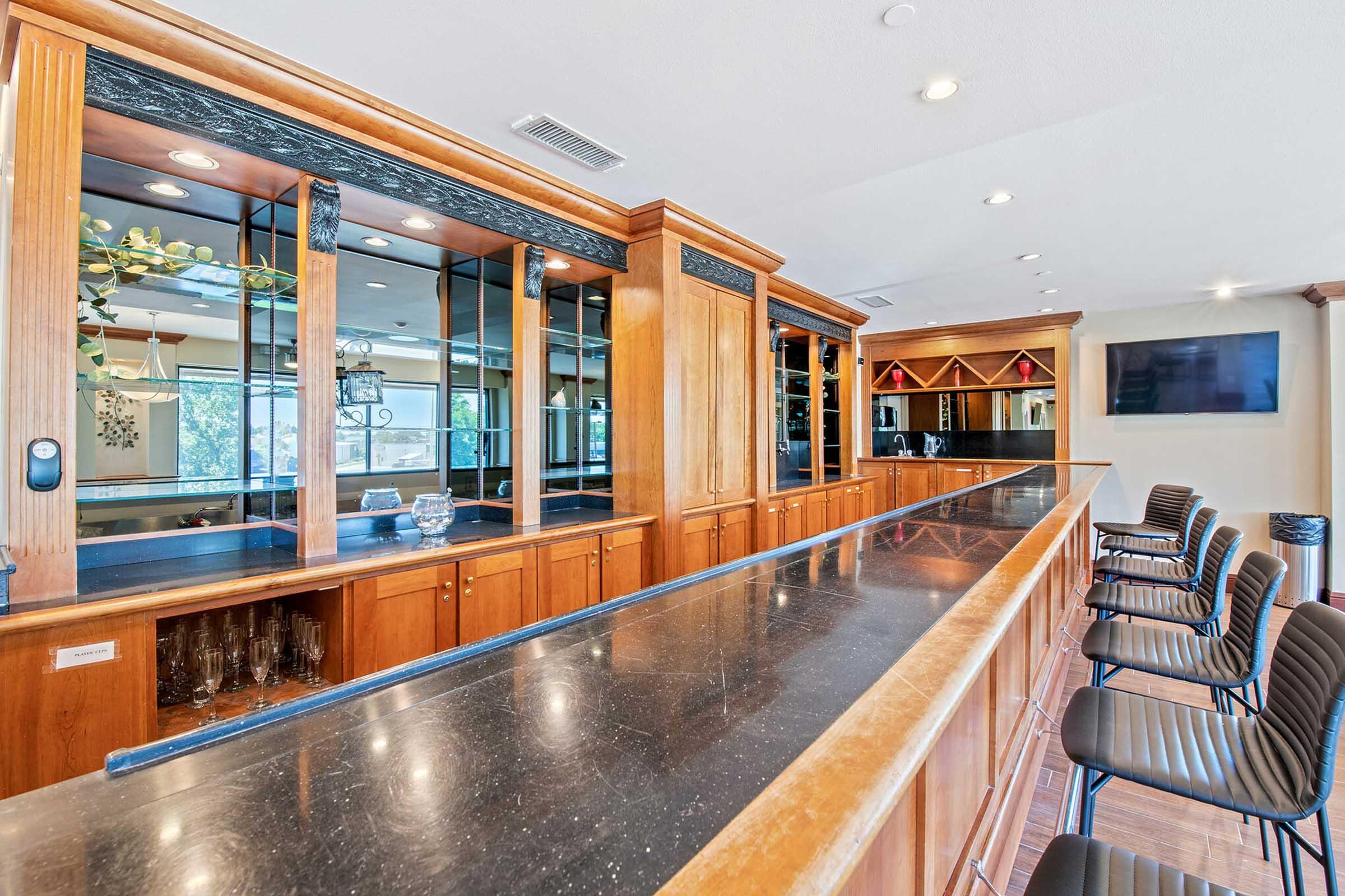 A modern bar area featuring polished wooden cabinetry and a granite countertop. The bar is lined with high-backed black stools, and there are mirrors and decorative elements on the walls. Soft lighting enhances the inviting atmosphere, creating a cozy space for socializing.