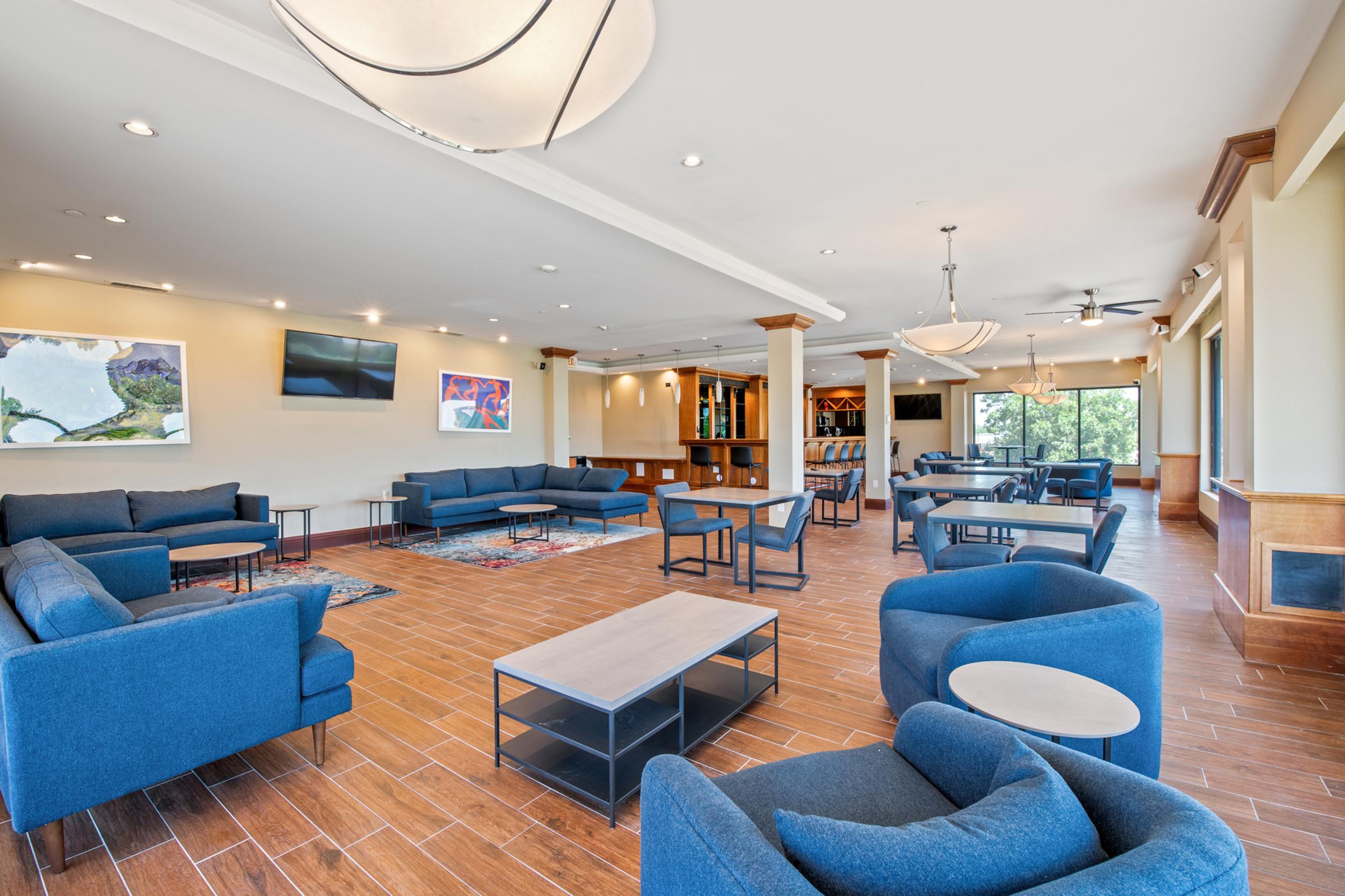 Modern lounge area featuring blue upholstered seating, coffee tables, and wooden flooring. Large windows provide natural light, and artwork decorates the walls. The space includes a built-in bookshelf and television, creating a comfortable and inviting atmosphere for relaxation or socializing.