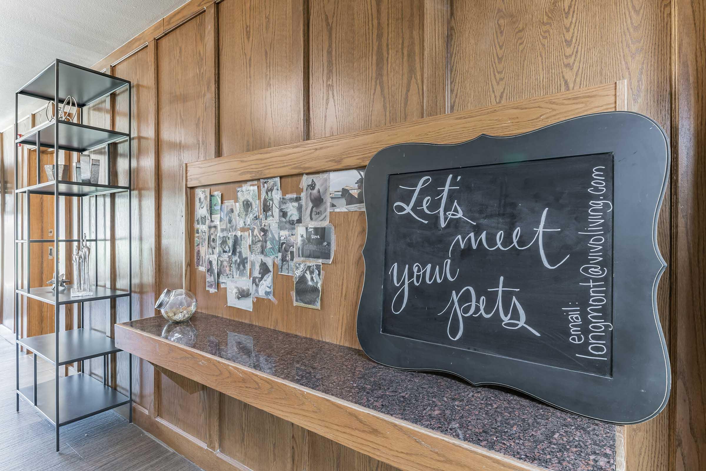 A wooden-paneled room with a modern black shelf holding decorative items. A large black chalkboard displays the text "Let's meet your pets" with an email address below. Surrounding the chalkboard are black-and-white photos pinned to the wall, creating a cozy, welcoming atmosphere.