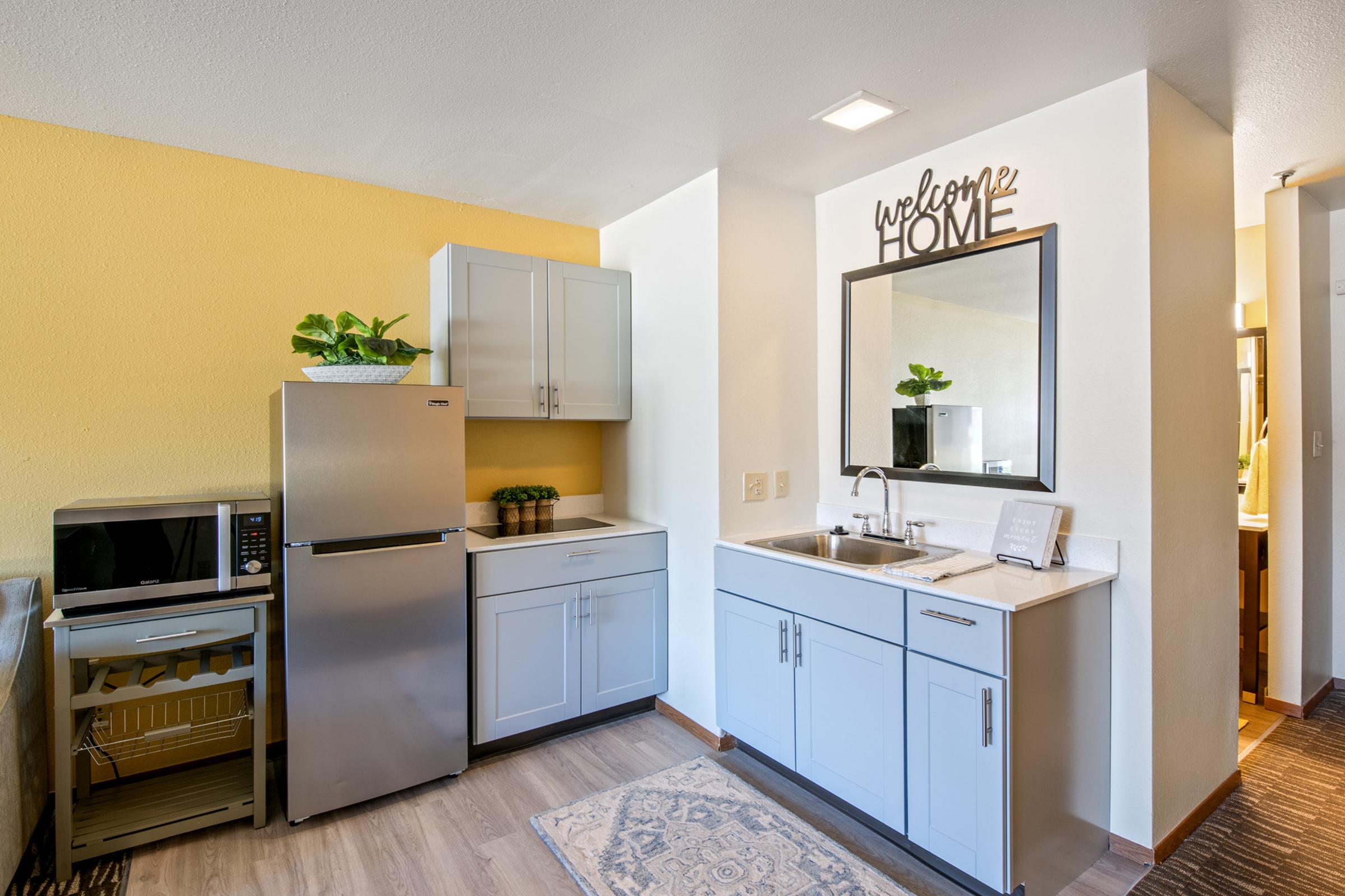 A modern kitchen featuring a stainless steel refrigerator, a microwave, and a sink with cabinets underneath. The walls are painted yellow and light gray, with a decorative "welcome HOME" sign above the sink. Small potted plants sit on the countertop, enhancing the cozy ambiance.