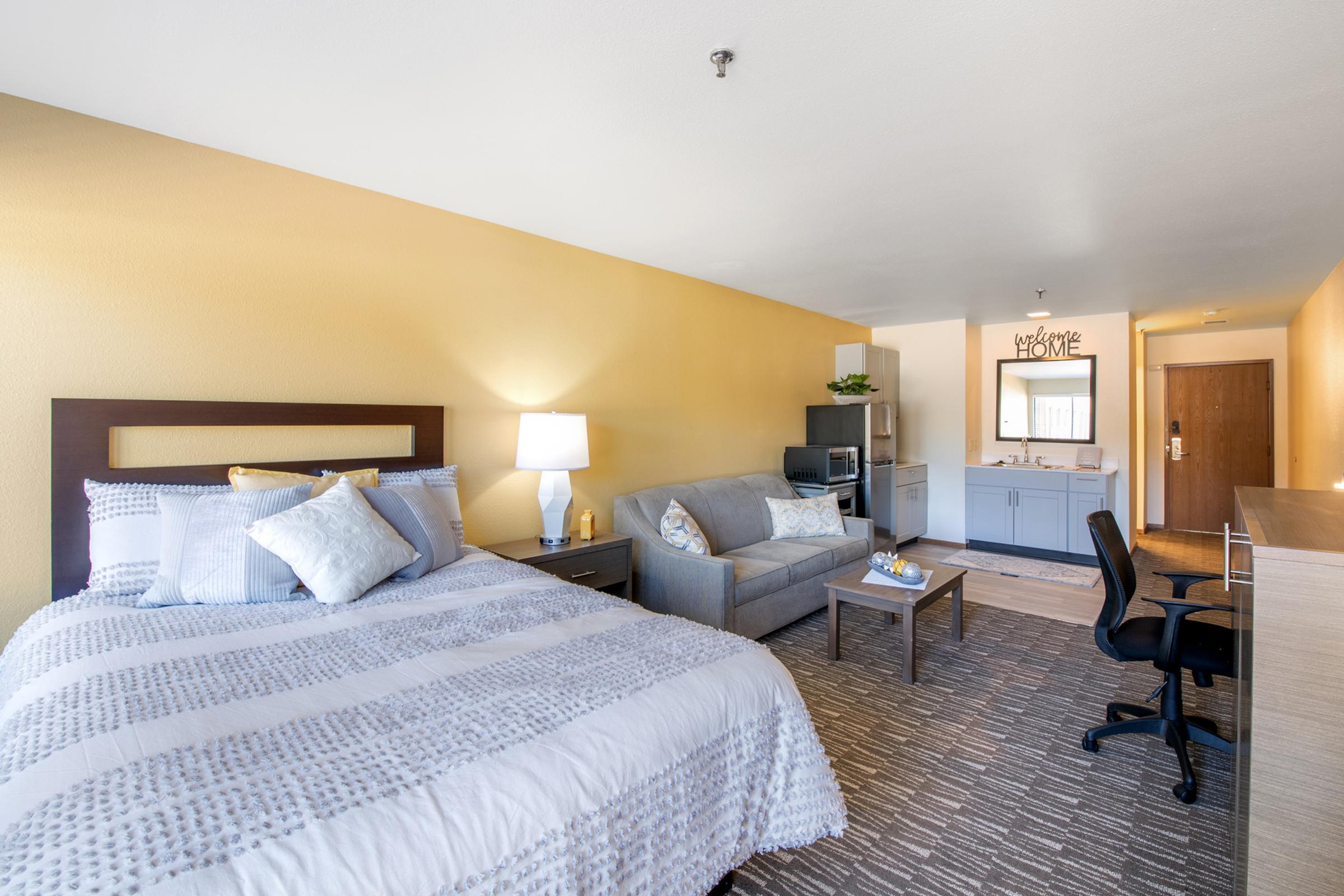 A cozy hotel room featuring a king-sized bed with decorative pillows, a gray couch, a small kitchenette with a sink and mini-fridge, a desk with a black chair, and a welcoming atmosphere created by warm yellow walls and soft lighting. A mirror and welcome message add a personal touch.