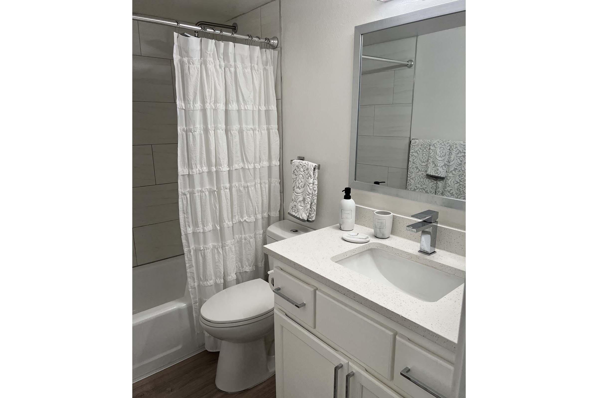 A clean and modern bathroom featuring a white shower curtain, a toilet, and a sink with a countertop. The sink has a soap dispenser and decorative towel. A large mirror is above the sink, and there are light-colored tiles on the walls and floor, creating a bright and spacious atmosphere.