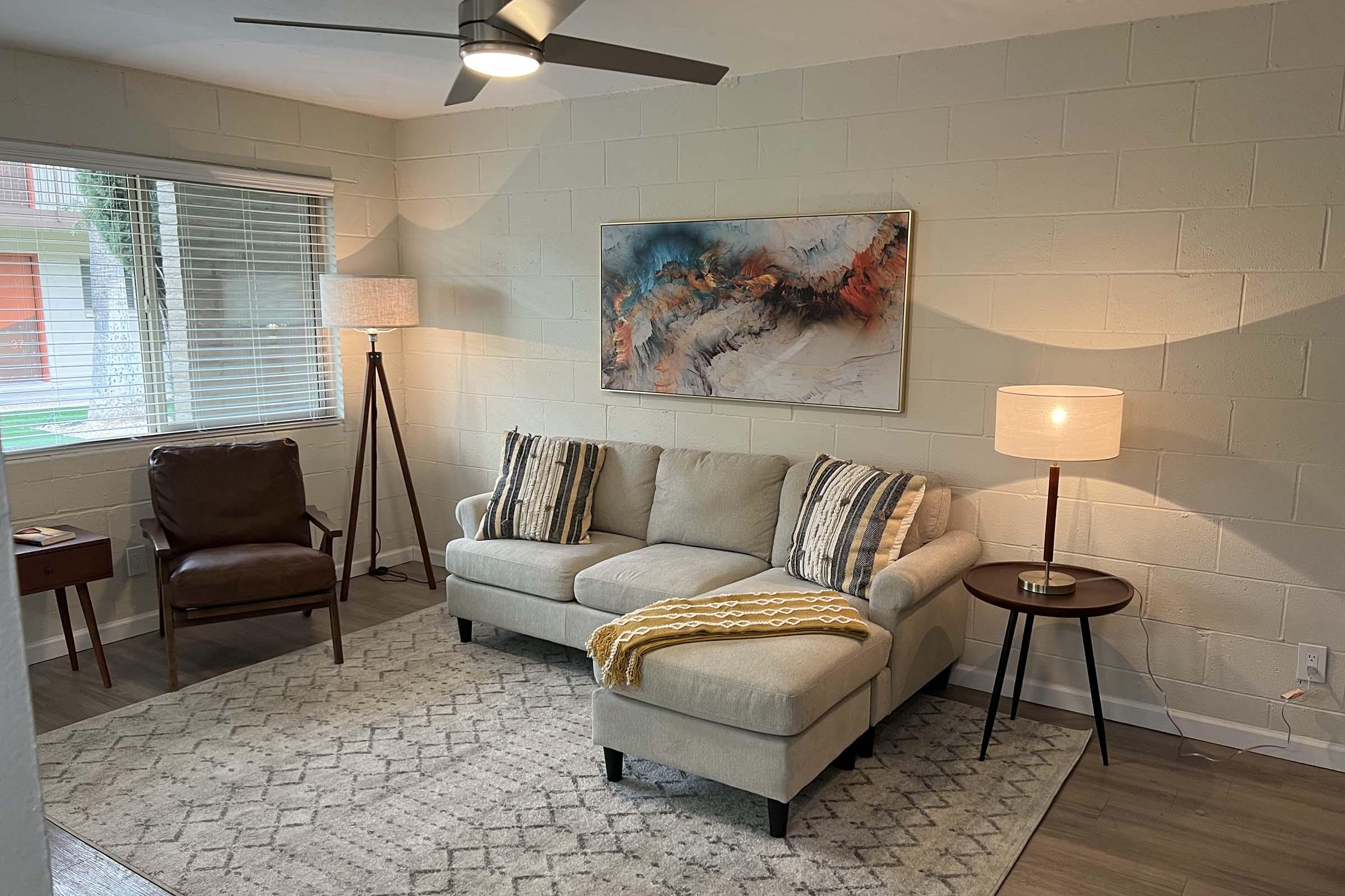 A cozy living room featuring a light-colored sectional sofa with patterned cushions, a warm yellow throw blanket, a brown leather armchair, and a wooden side table. A floor lamp and a table lamp provide soft lighting, while a large abstract painting adorns the wall above the sofa. Natural light comes in from the window.