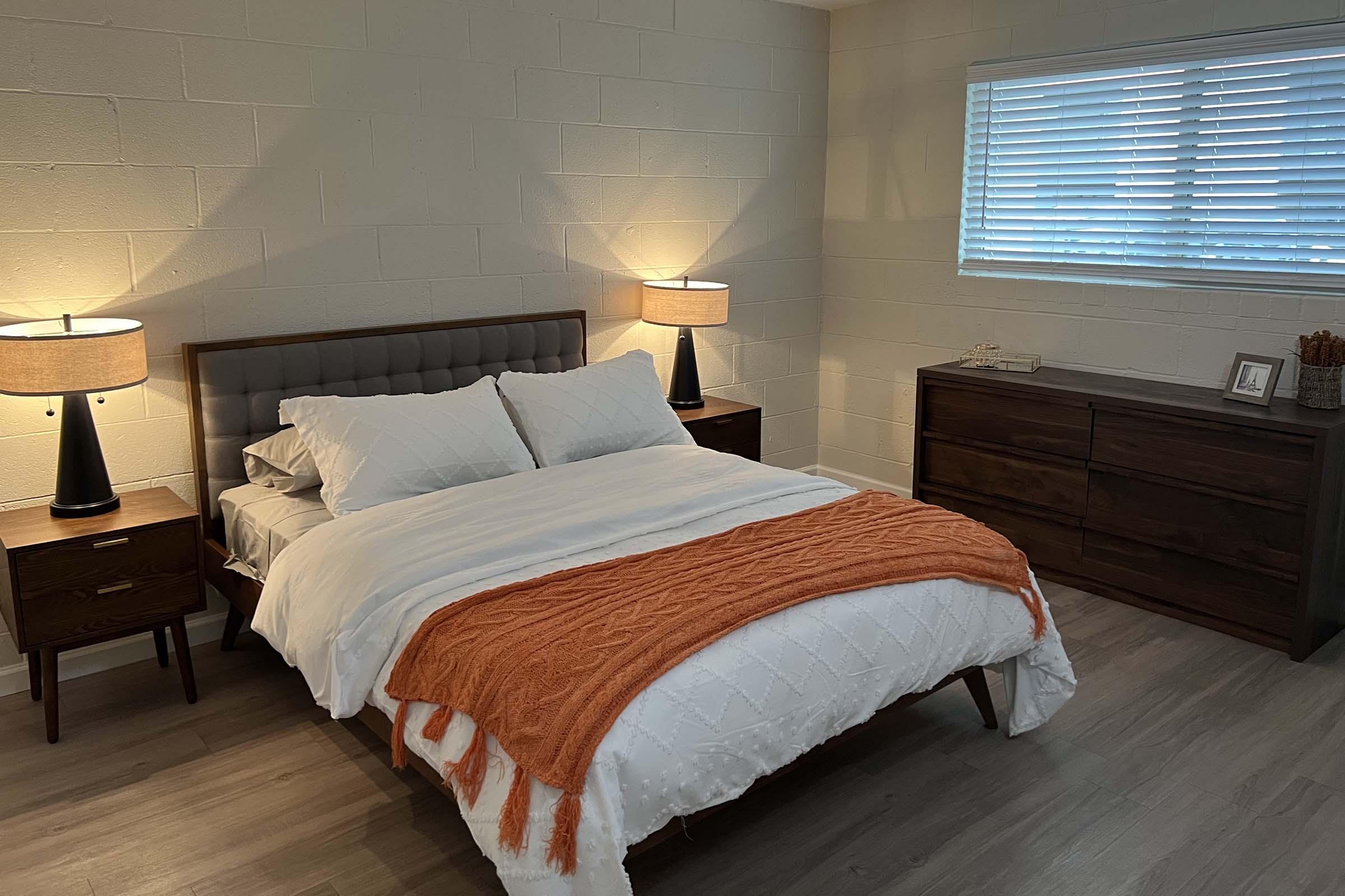 A cozy bedroom featuring a stylish bed with white linens and an orange throw blanket. There are two bedside tables with lamps, and a wooden dresser against the wall. Natural light filters through a window with white blinds, creating a warm and inviting atmosphere.