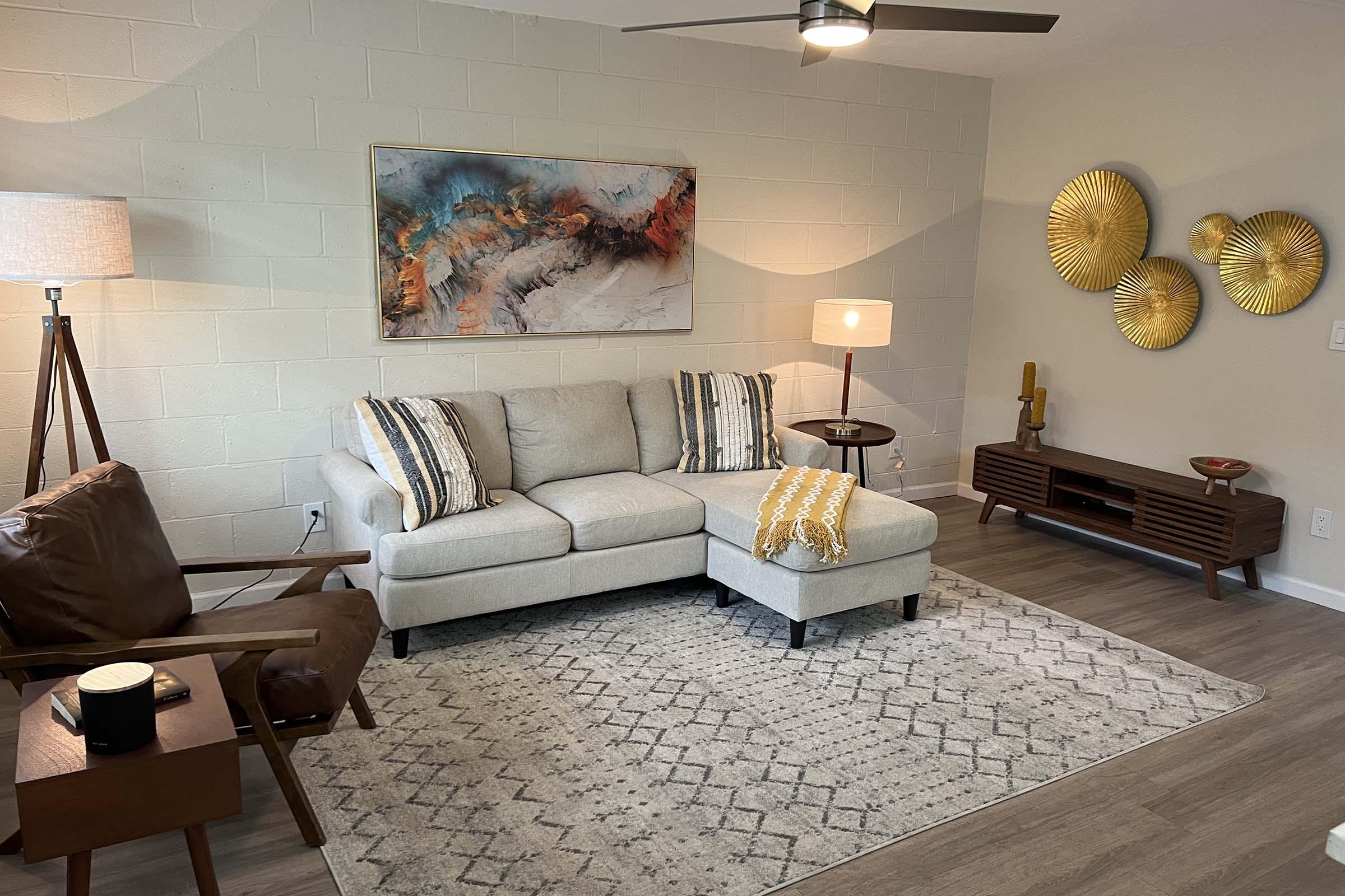 A modern living room featuring a light-colored sectional sofa with decorative pillows, an accent chair, a coffee table, and a wall-mounted artwork. The space includes a floor lamp and a side table with a lamp, along with a textured area rug and a media console with circular wall decor. Natural light enhances the cozy atmosphere.