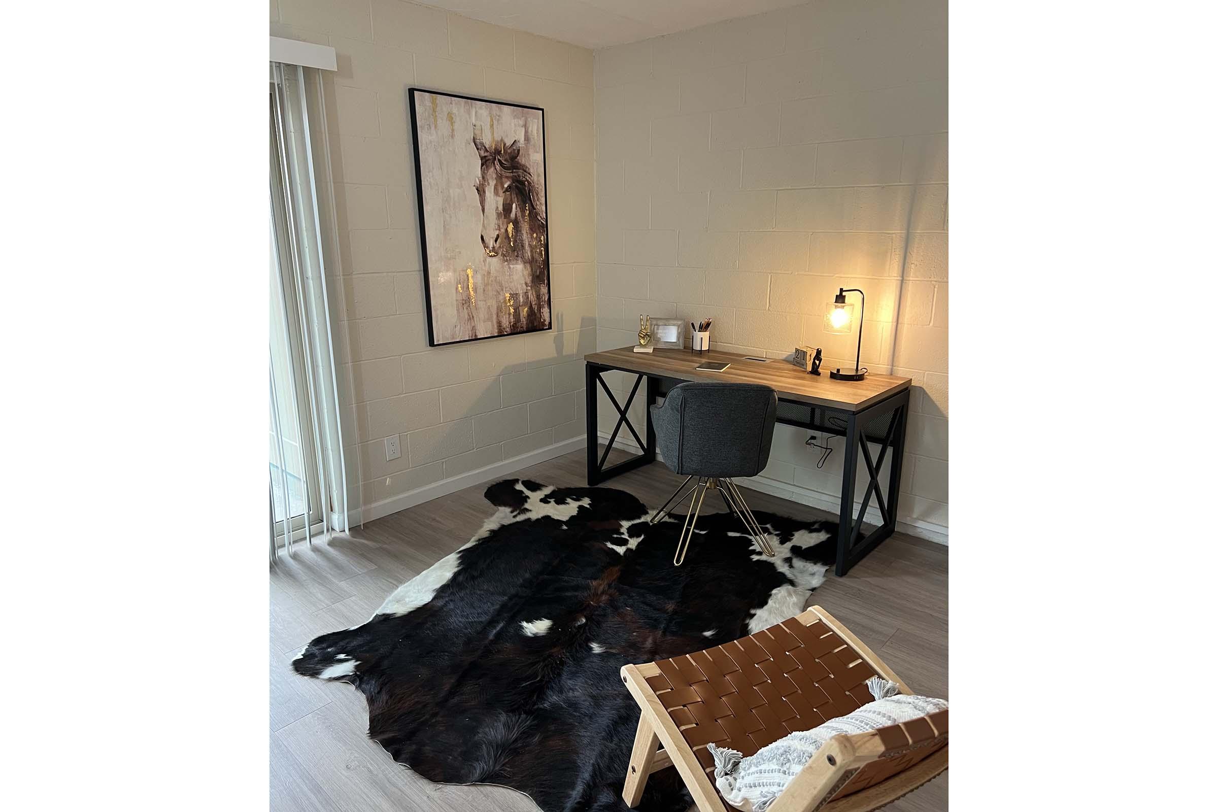A modern workspace featuring a wooden desk with a lamp, a black chair, and a decorative wall art of a horse. A cowhide rug is positioned on the floor, complementing the sleek decor. Natural light streams in through sliding glass doors, creating a warm and inviting atmosphere.
