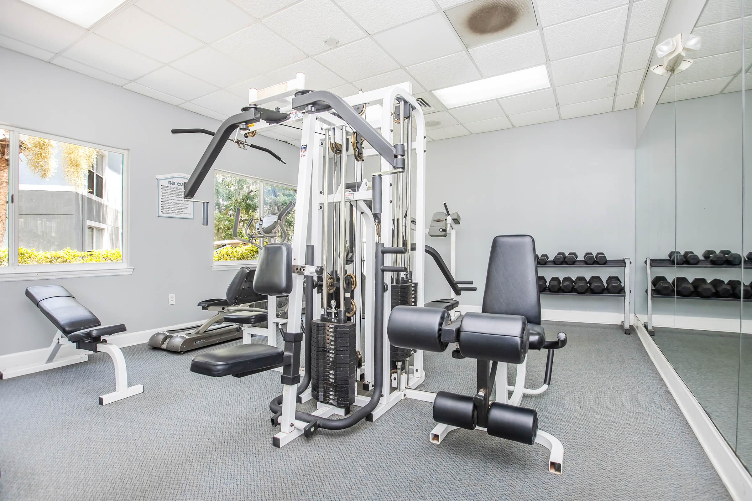 A well-equipped gym interior featuring a multi-gym machine, a bench, and a set of dumbbells. The room has bright lighting, a mirror on one wall, and large windows allowing natural light to enter. The floor is carpeted, creating a comfortable workout environment.