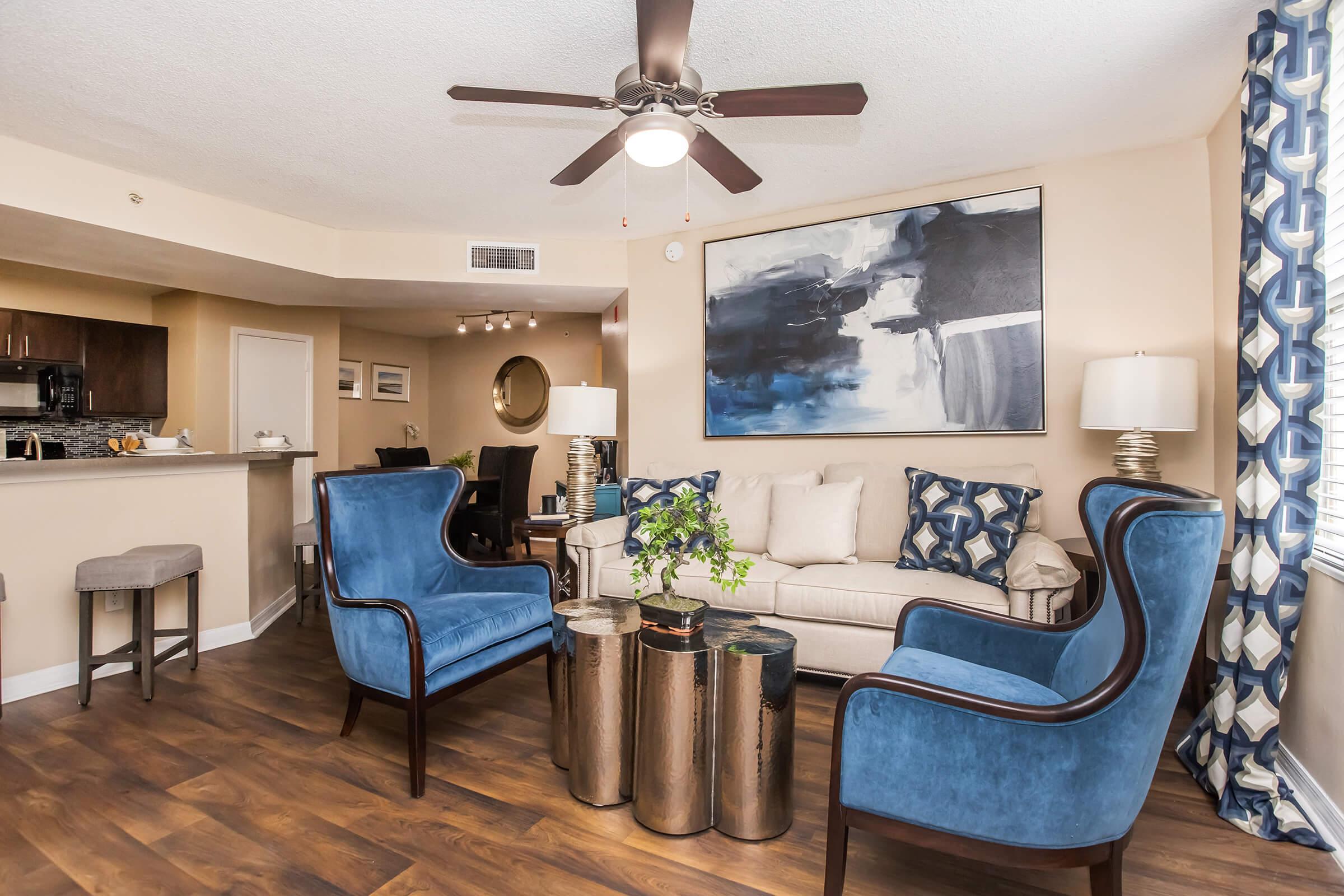 A modern living room featuring a light-colored sofa with decorative pillows, two blue accent chairs, and a coffee table made of metallic stools. The room has a ceiling fan, a large abstract painting on the wall, and stylish curtains. A small bar area with stools is visible in the background.