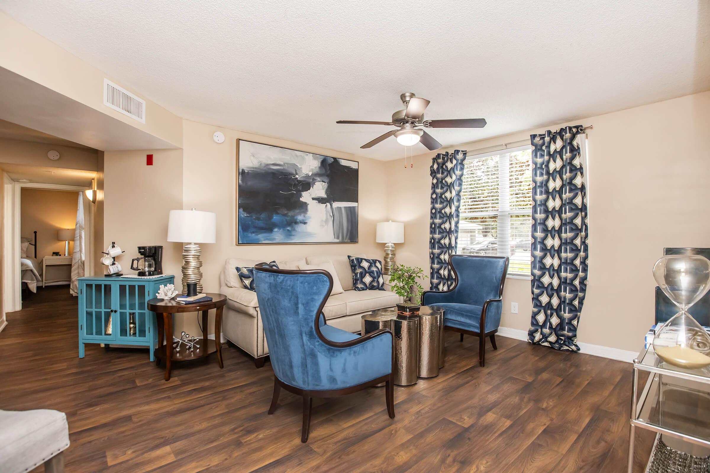 A modern living room featuring a beige sofa and two blue accent chairs, a ceiling fan, and a stylish coffee table. There are decorative elements, including a potted plant and artwork on the wall. Large windows with blue patterned curtains allow natural light, and the flooring is wooden.